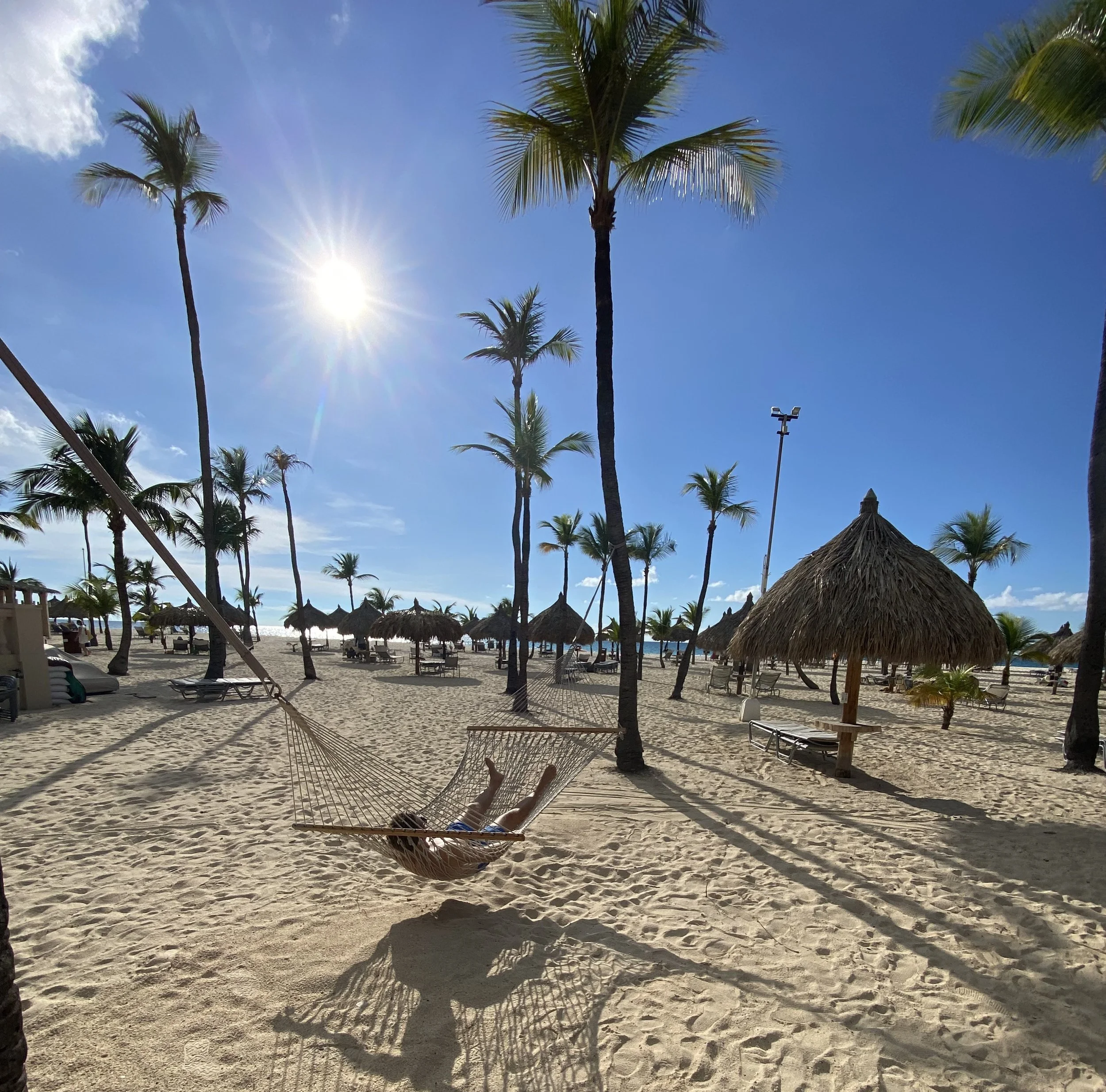 Person ligt in een hangmat op een zonnig strand met palmbomen en strandhutten.