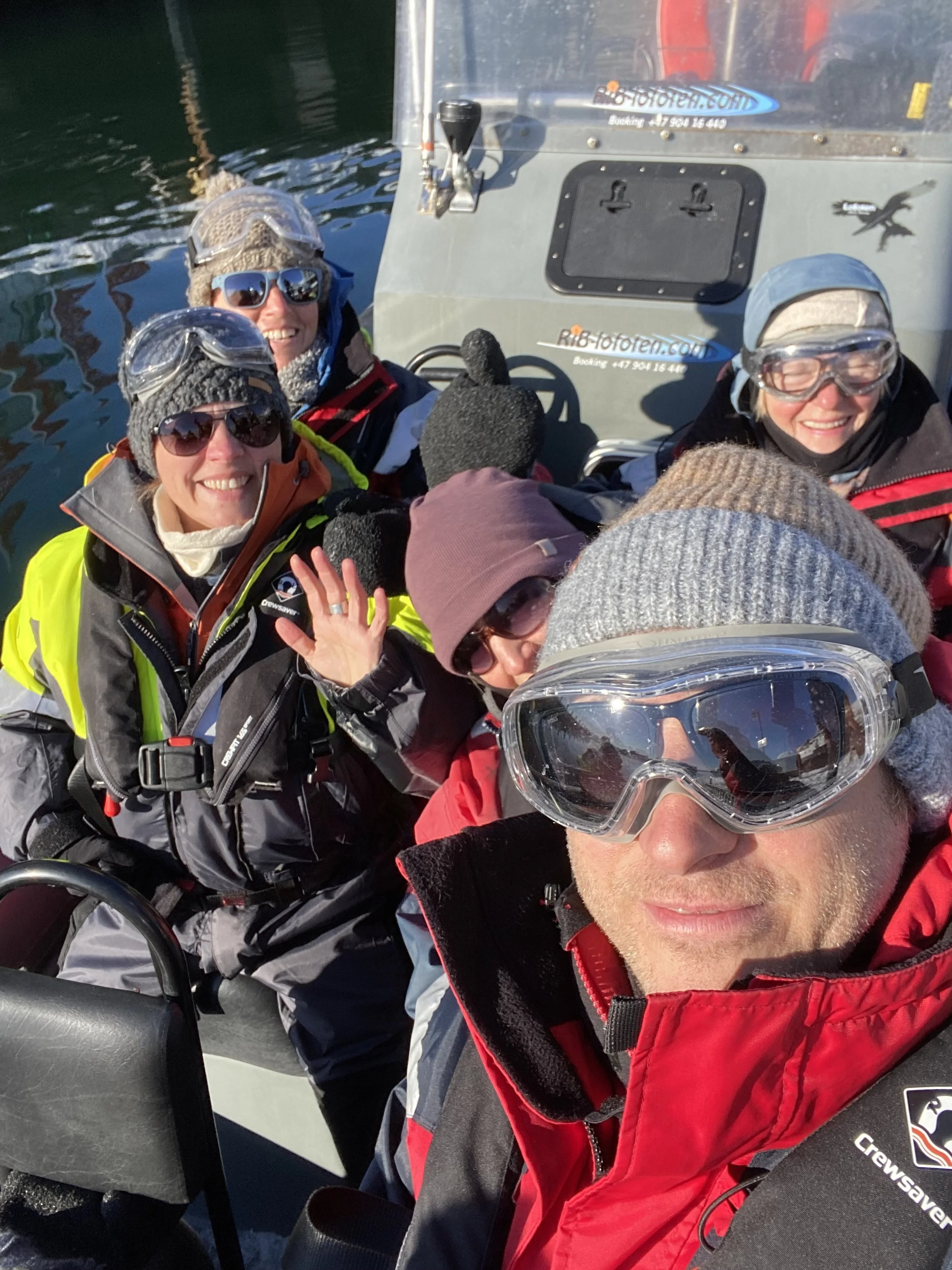 Vijf mensen gekleed in winterkleding en skibrillen, vrolijk op een boot op het water.