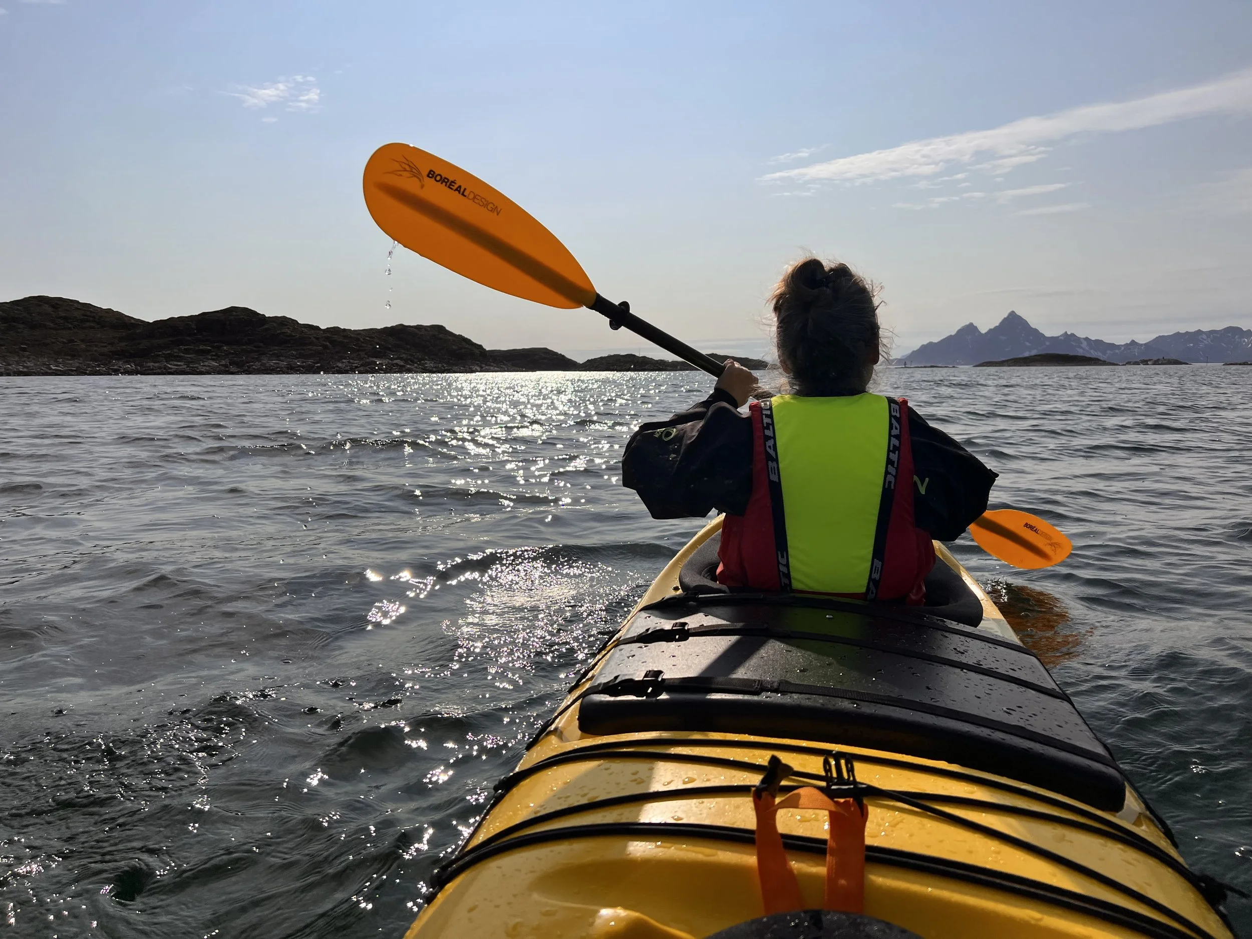 Persoon kajakt met omgeving van water, kleine eilanden en bergen op de achtergrond, onder een lichte bewolkte hemel.