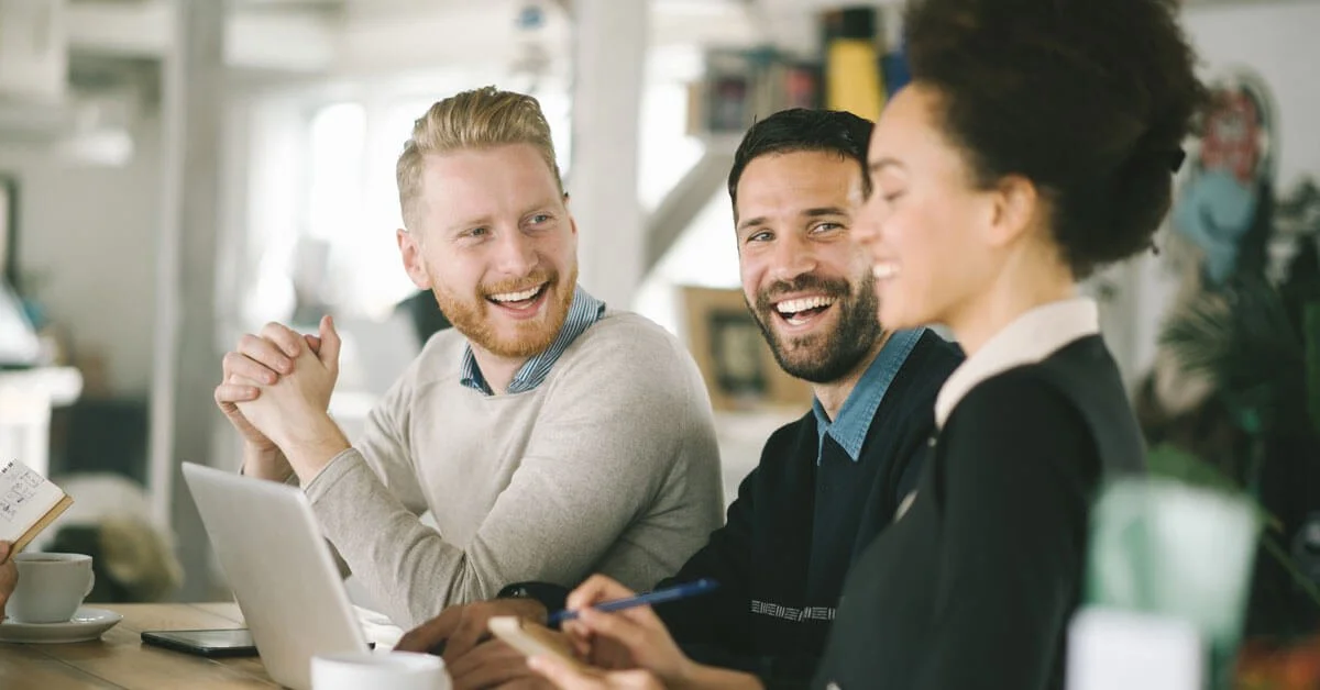 Drie lachende mensen in een kantooromgeving, genietend van een gesprek.