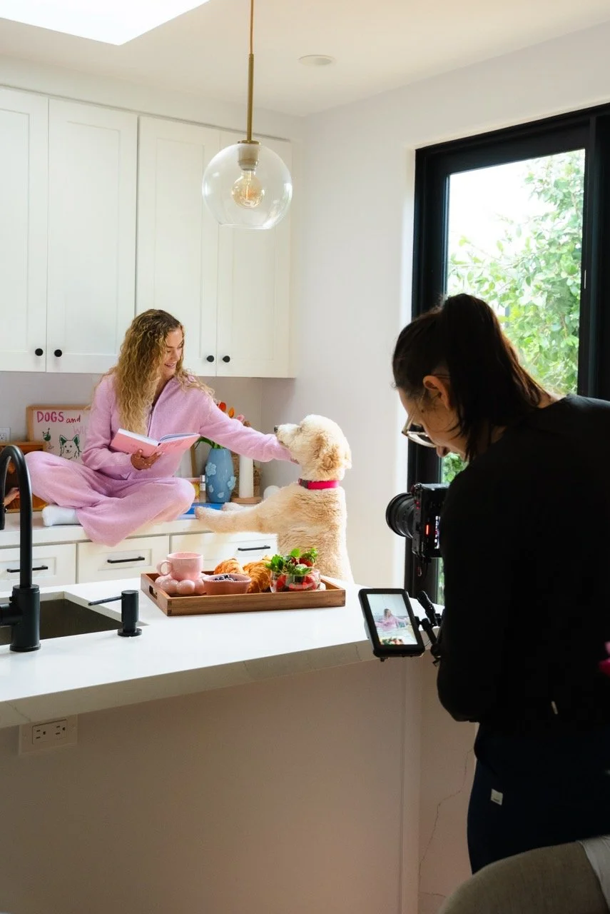 A woman in pink pajamas sitting on a kitchen counter, petting a white poodle dog, with another woman filming the scene with a camera.