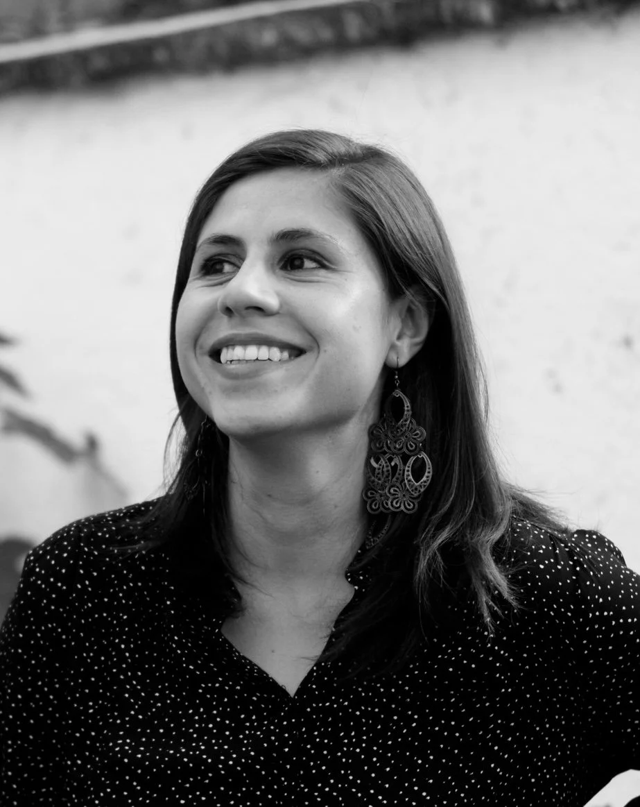 Retrato en blanco y negro de una mujer sonriendo, con cabello largo y pendientes grandes, llevando blusa de puntos blancos en un fondo difuso.
