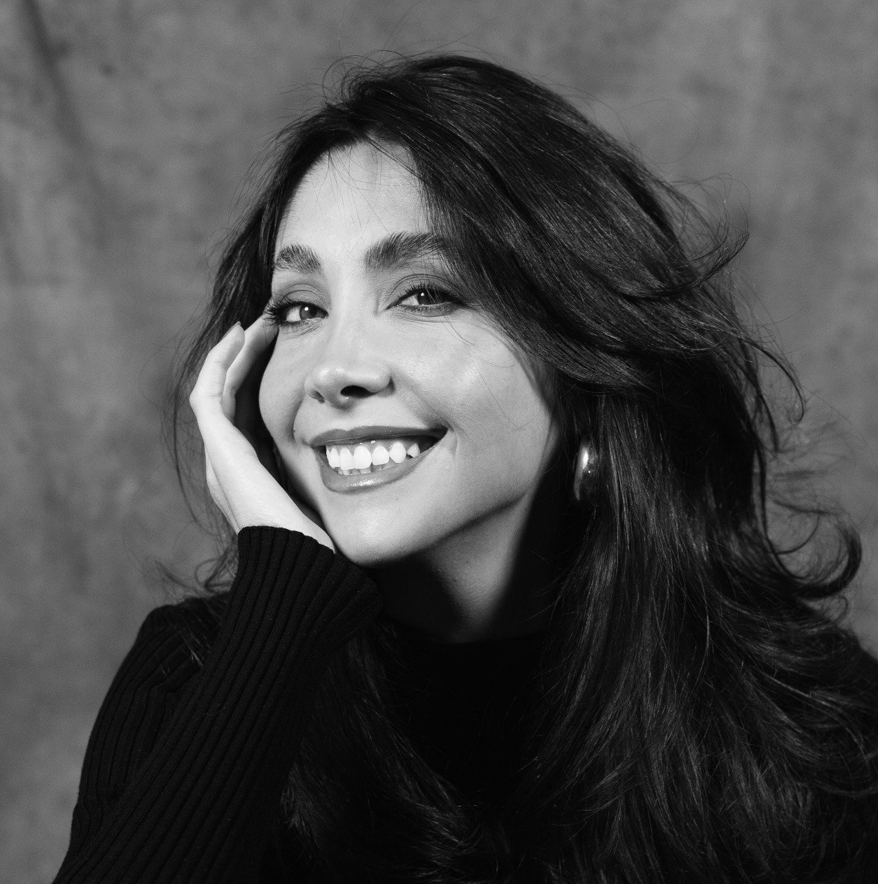 Retrato en blanco y negro de una mujer sonriendo, con cabello largo y rizado, usando un suéter de manga larga y un pendiente grande.