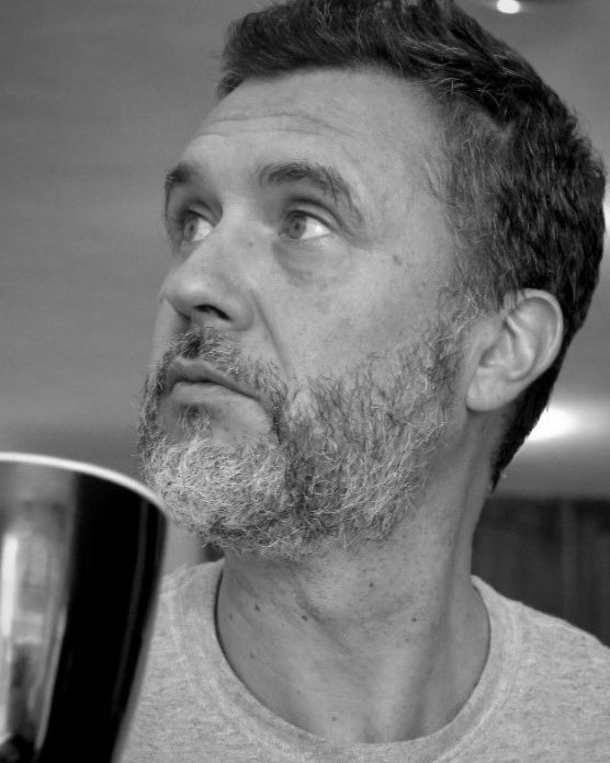 Hombre de cabello oscuro y barba canosa, mirada hacia arriba, en un entorno de interior, en escala de grises.