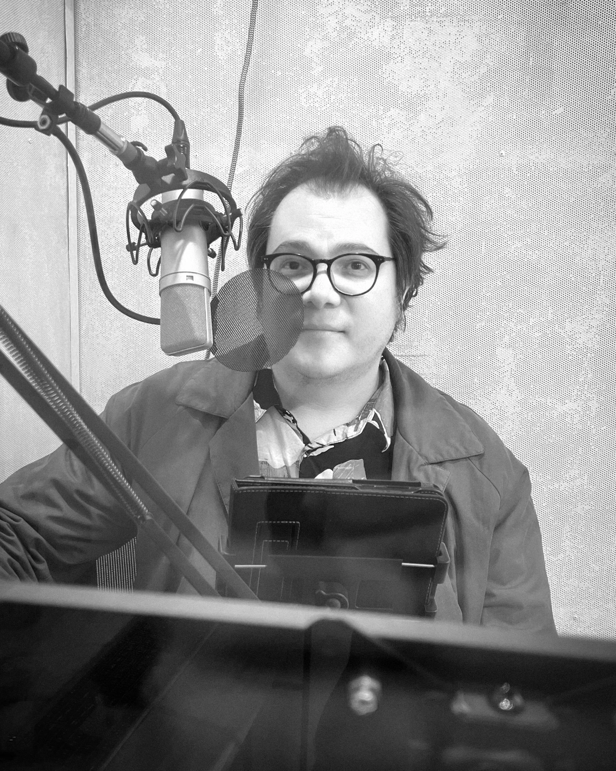 Un hombre con gafas y cabello despeinado sentado en un estudio de grabación, frente a un micrófono de estudio con filtro, con equipo de grabación y una tablet en la mesa.