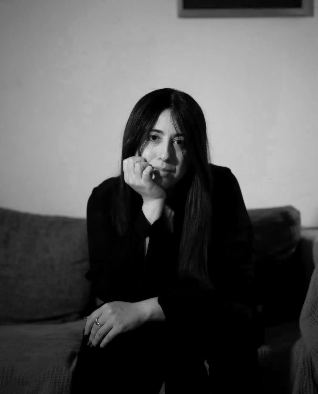 Retrato en blanco y negro de una mujer joven con cabello largo y oscuro, descansando su rostro en su mano, sentada en un sofá, en un ambiente interior