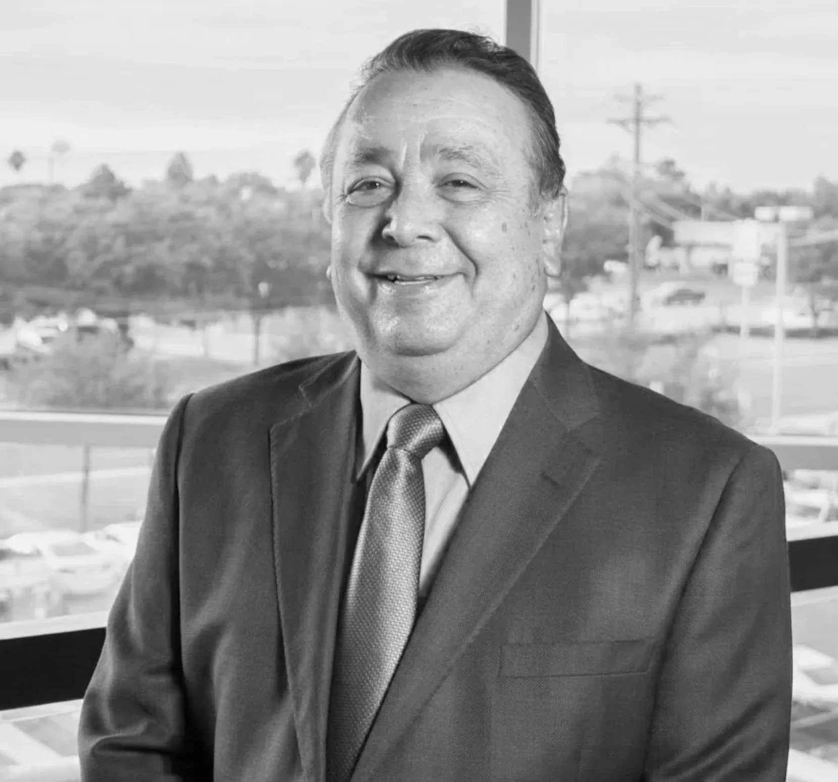 Hombre mayor sonriendo con traje y corbata, en una oficina con una ventana grande al fondo.