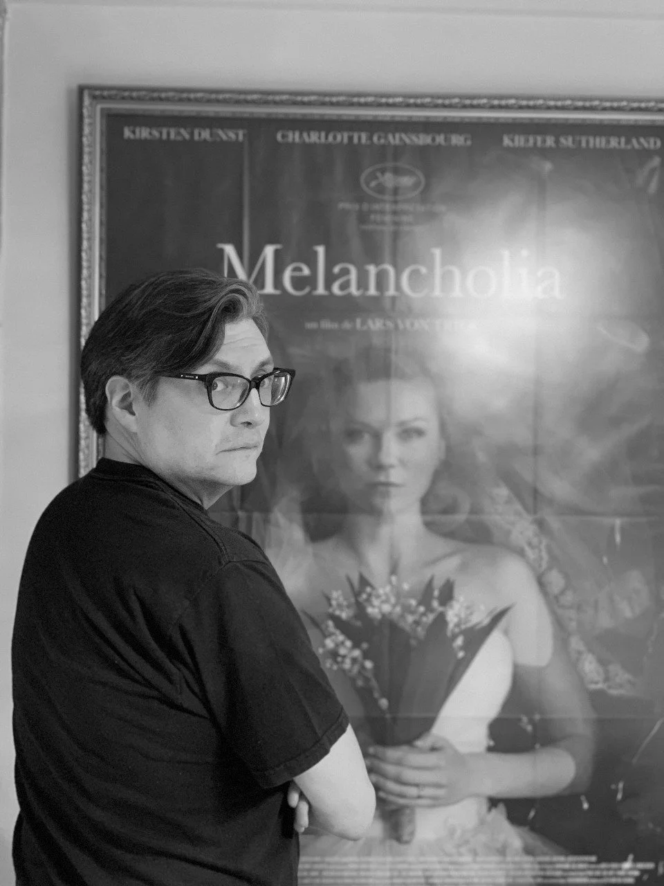 Hombre con gafas observa una cartelera de la película 'Melancholia' con imagen de una mujer sosteniendo un ramo de flores.