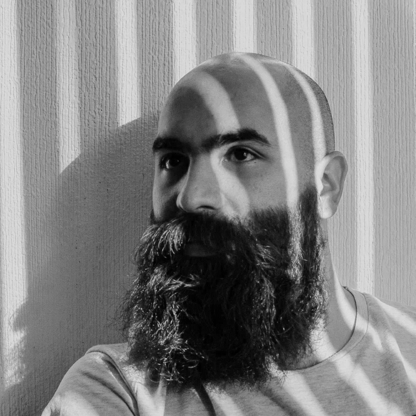 Hombre calvo con barba grande, sombras de persianas en la cara, fondo con textura de pared.