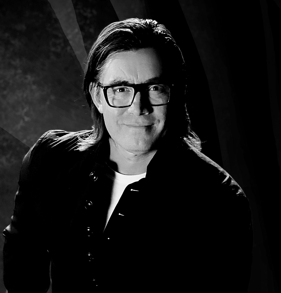 Retrato en blanco y negro de una persona con gafas y cabello largo, sonriendo discretamente, fondo oscuro.