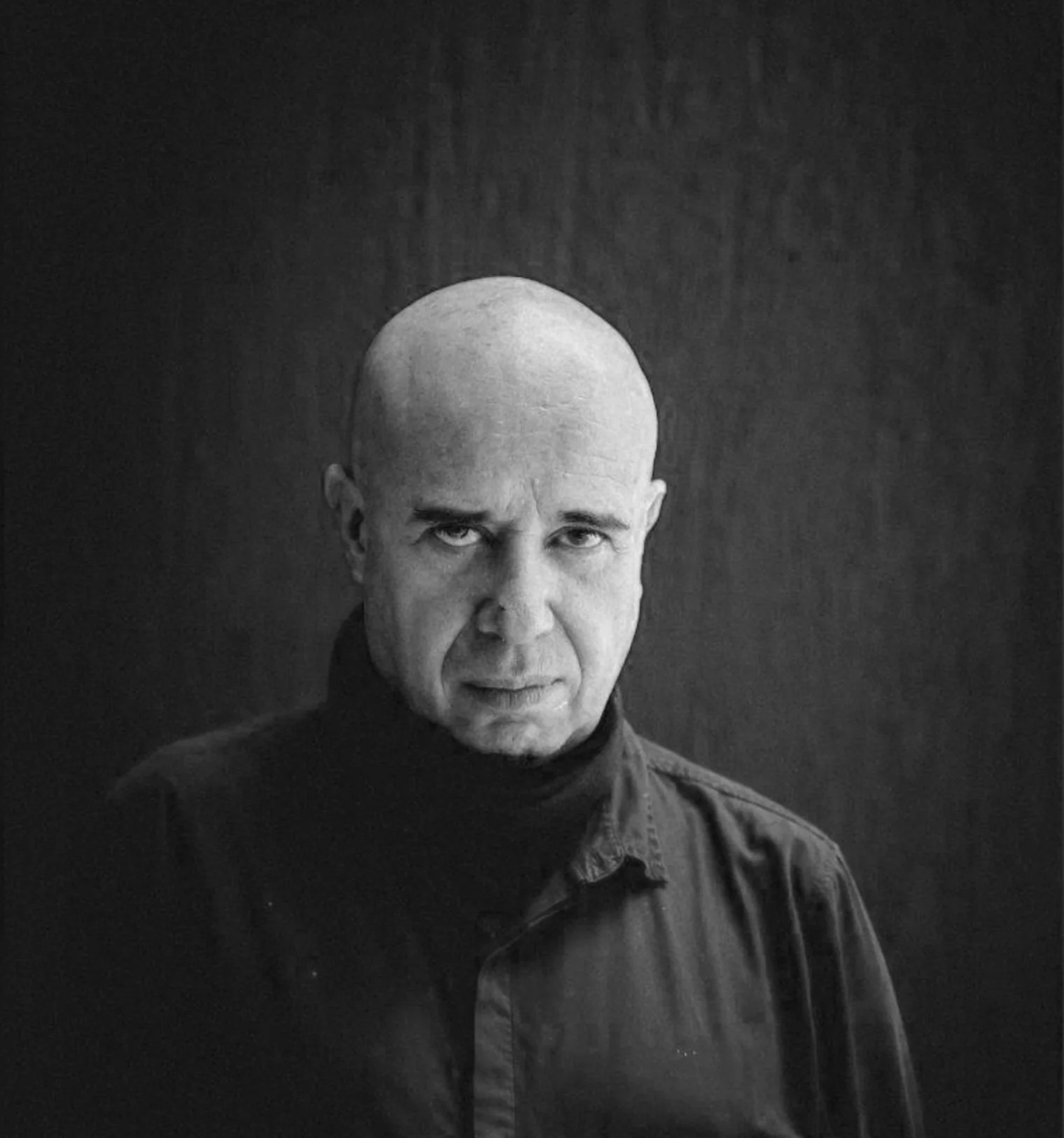 Retrato en blanco y negro de un hombre calvo con expresión seria, vistiendo una camisa oscura, frente a un fondo oscuro.