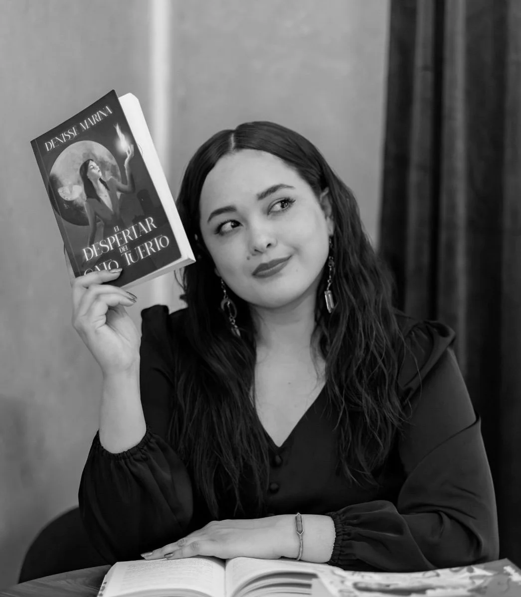 Retrato de una mujer con cabello largo y oscuro, con pendientes grandes, sosteniendo un libro titulado 'El disipar del casto juicio' de Delyssi Mariana, en color negro y blanco, en una mesa con un libro abierto.