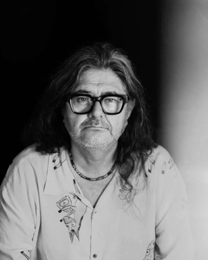 Retrato en blanco y negro de un hombre mayor con cabello largo, barba y bigote, lleva gafas oscuras y camiseta con dibujos, mirada seria.