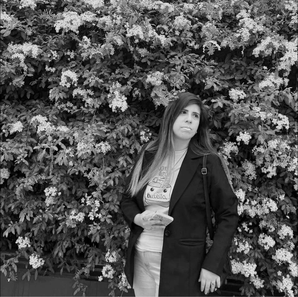 Mujer de pie frente a un arbusto con flores pequeñas, vistiendo un abrigo y sosteniendo un teléfono.