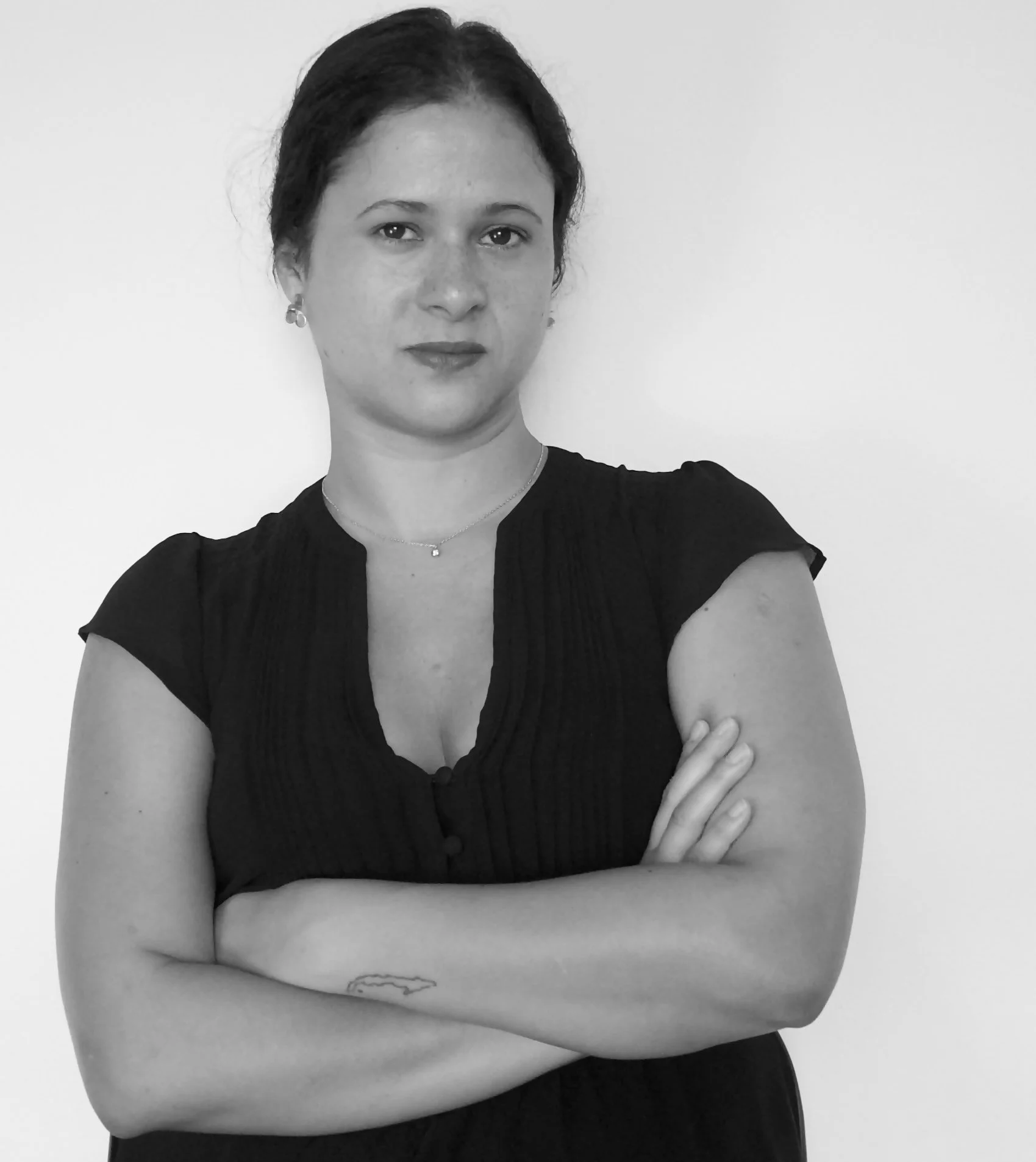 Mujer de cabello oscuro con los brazos cruzados, vistiendo una blusa oscura, en fondo blanco. Imagen en blanco y negro.