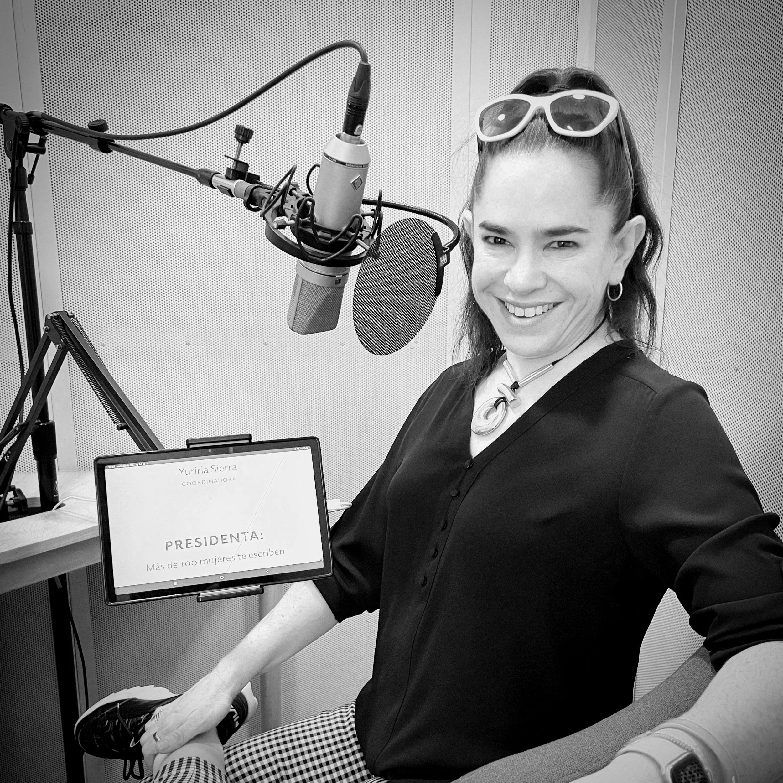 Mujer sonriendo frente a un micrófono de estudio, con gafas de sol en la cabeza. Hay una tableta a su lado con texto que dice 'Más de 100 mujeres te escriben'. El ambiente es un estudio de grabación.