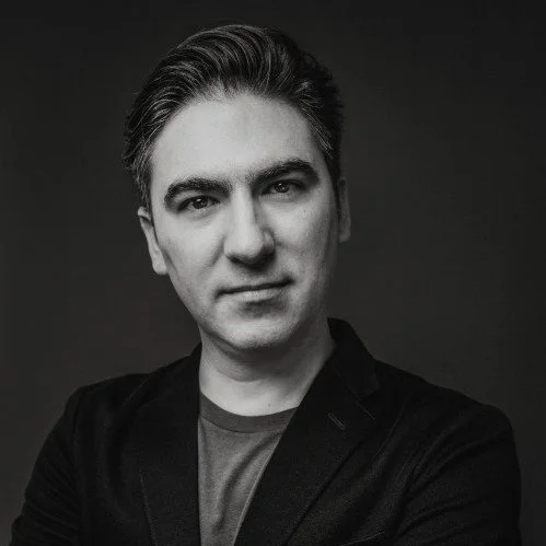 Retrato en blanco y negro de un hombre joven con cabello oscuro peinado hacia atrás, usando una chaqueta oscura y camiseta gris, frente a un fondo gris oscuro.