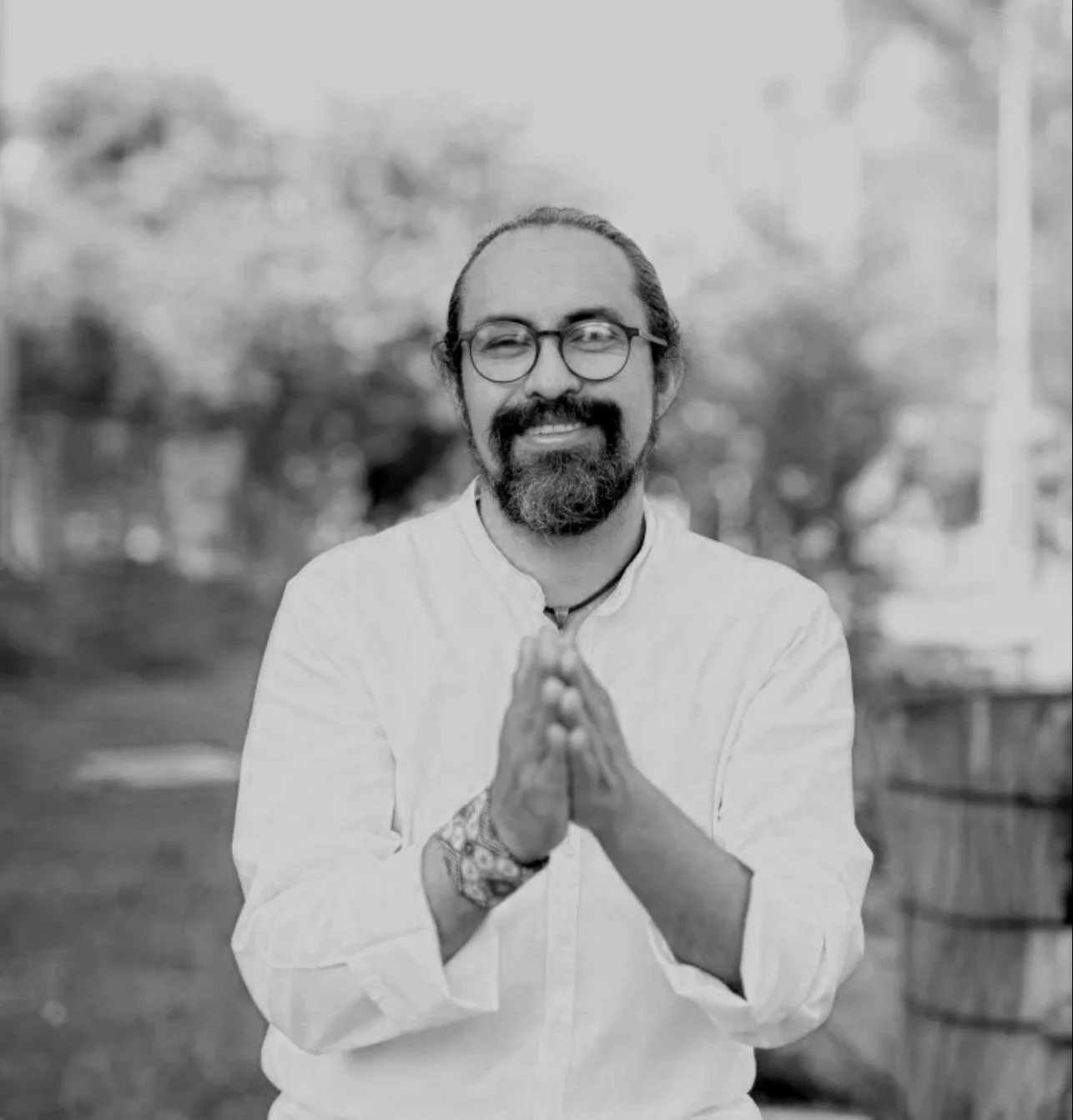 Hombre con gafas y barba sonríe, lleva camisa blanca y pulsera de patrón, fondo difuminado, manos juntas frente al pecho.