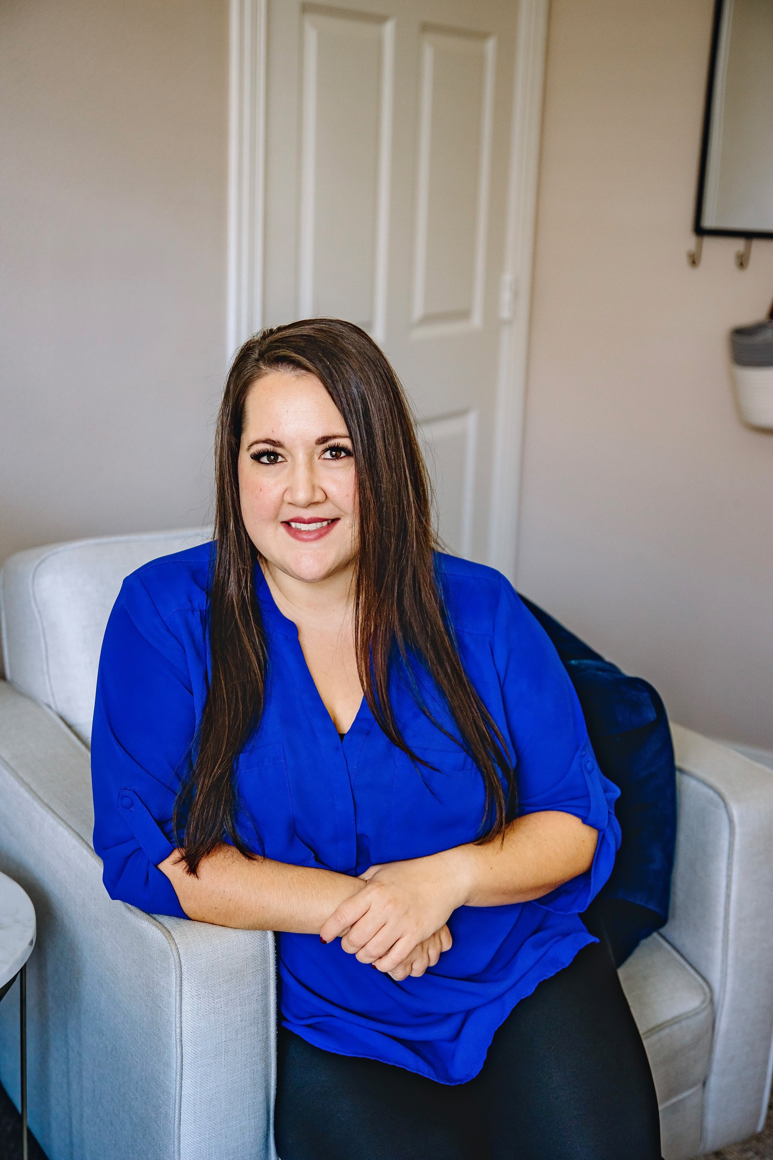 A woman with long brown hair wearing a blue top, sitting on a beige chair in a room with beige walls, a closed white door, and a wall-mounted white device.