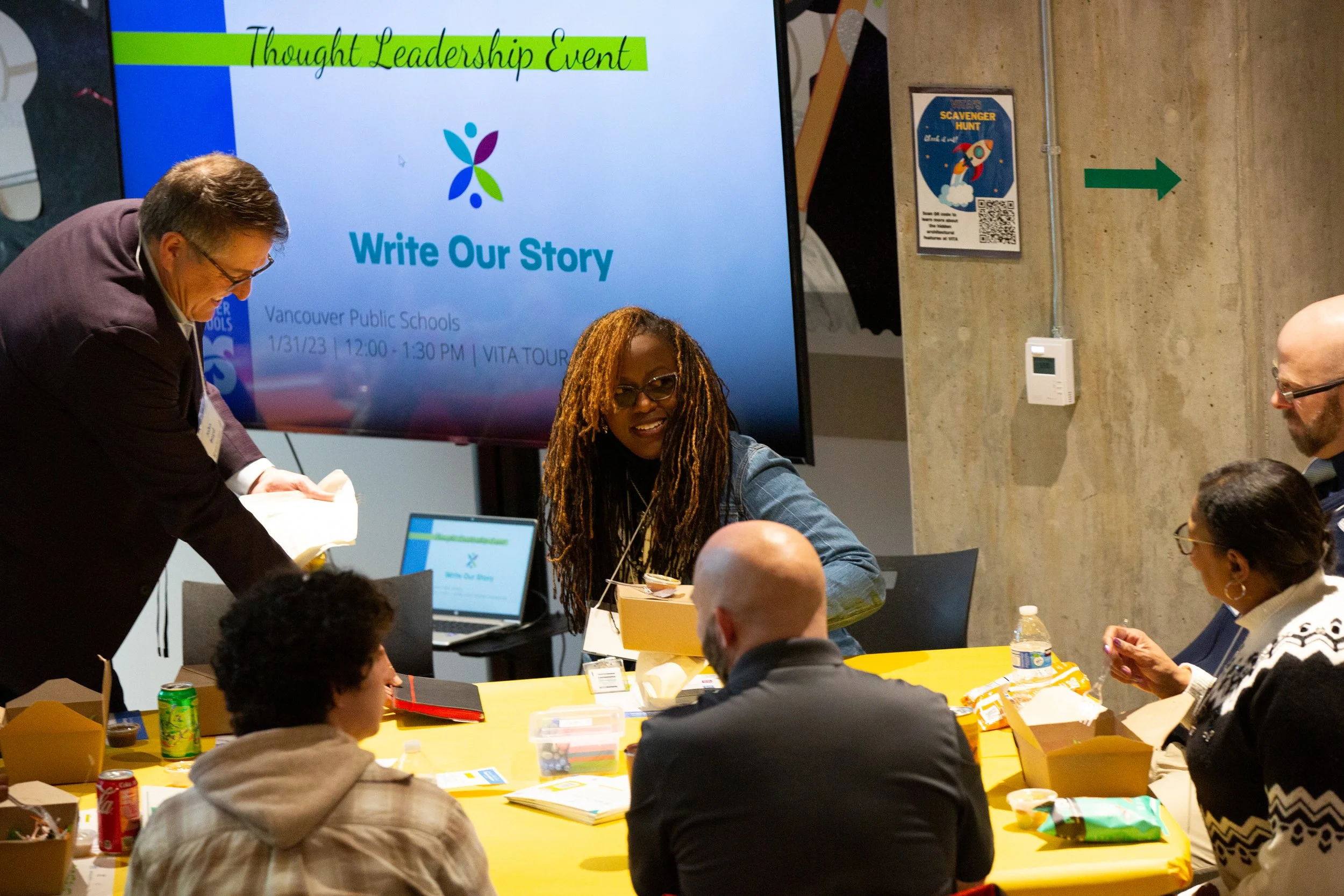 A group of people sitting around a table at a workshop, talking together. There is a slide behind them with the Write Our Story logo and the words "Thought Leadership Event"
