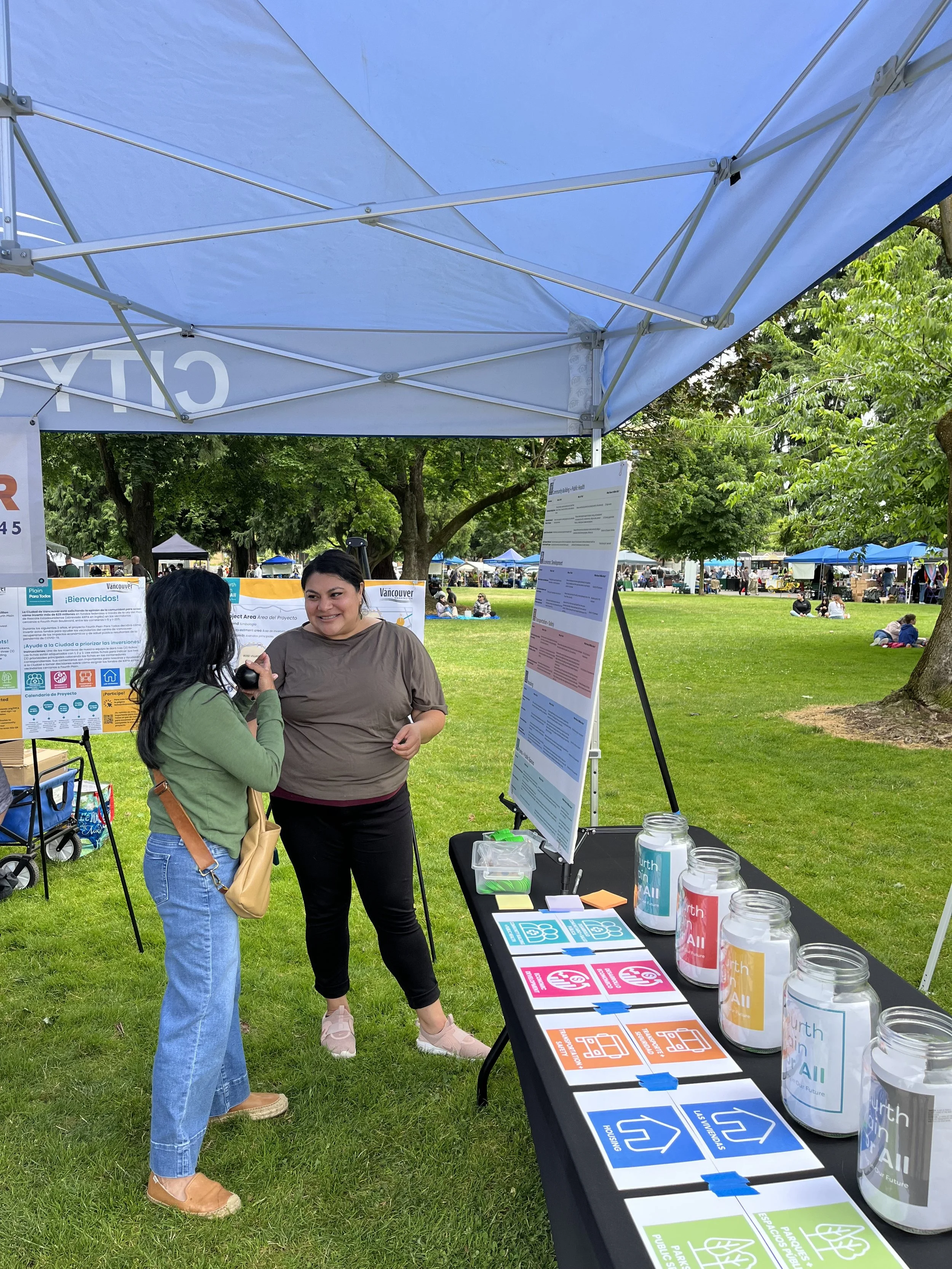 An image of a woman in a brown shirt and black pants engaging with a woman in a green shirt and blue pants at an outdoor event in front of a Fourth Plain for All table and supporting display boards