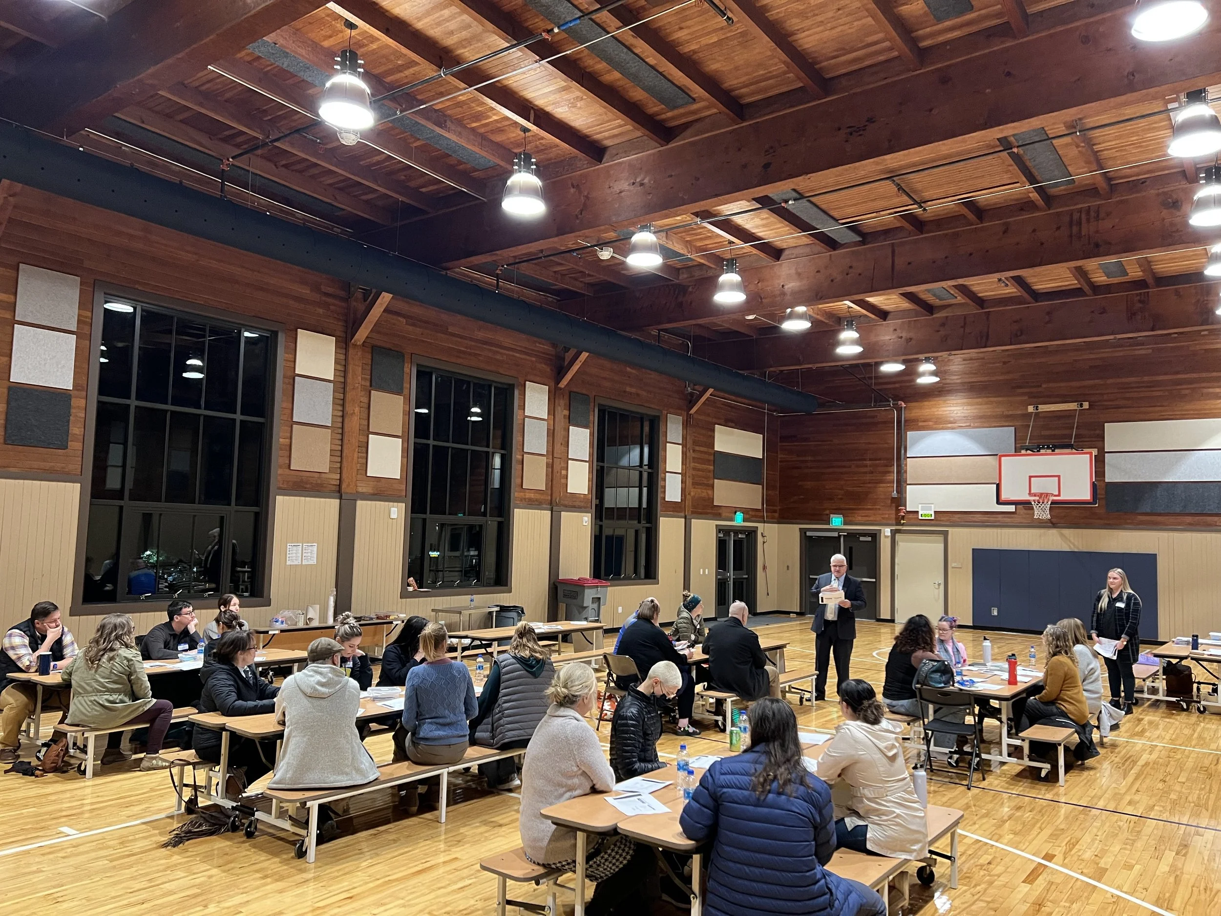 Two people standing in front of a group of people sitting at desks in a gymnasium