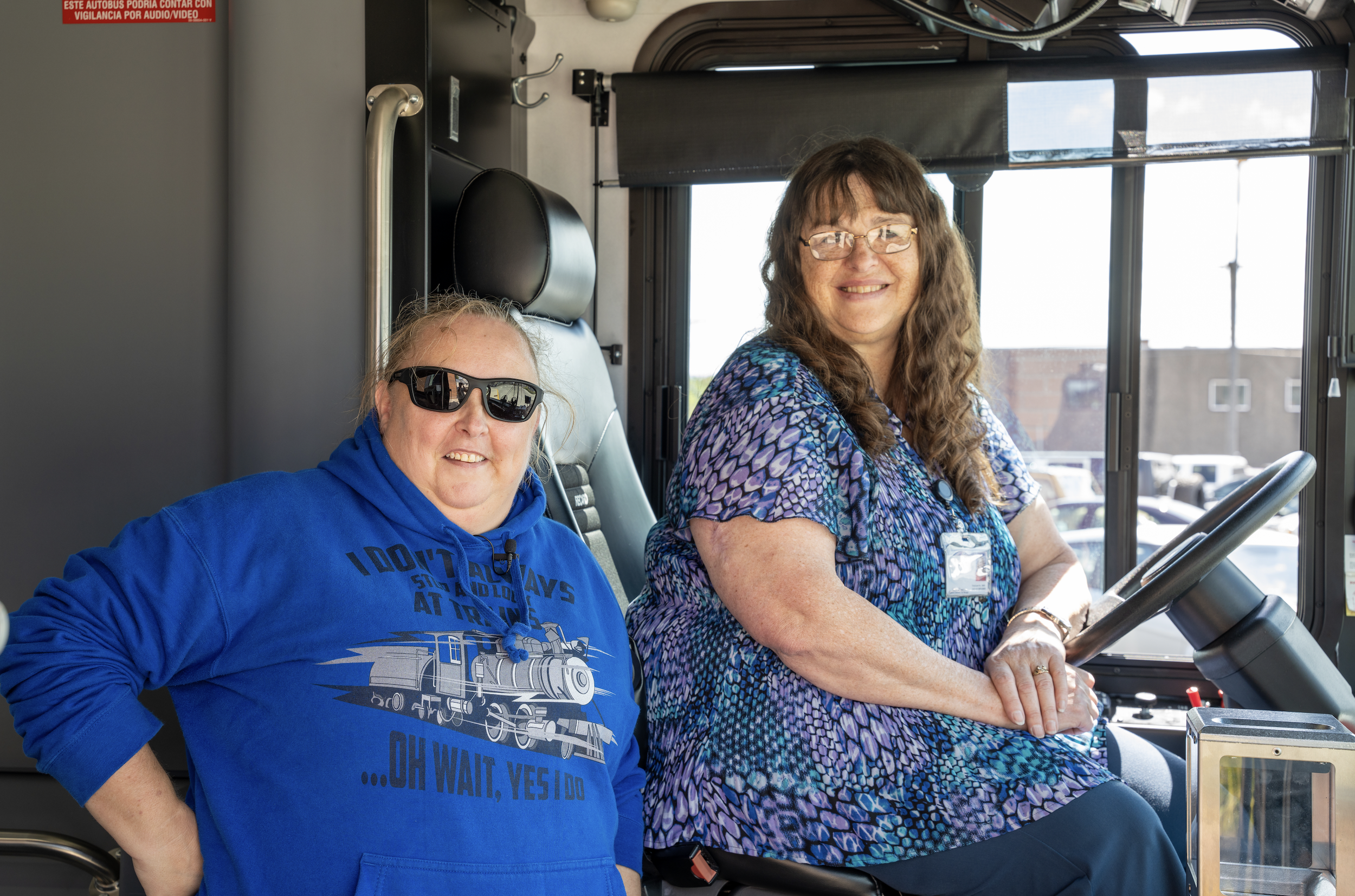 Woman sitting in the drivers seat in a bus with a second woman standing next to her smiling