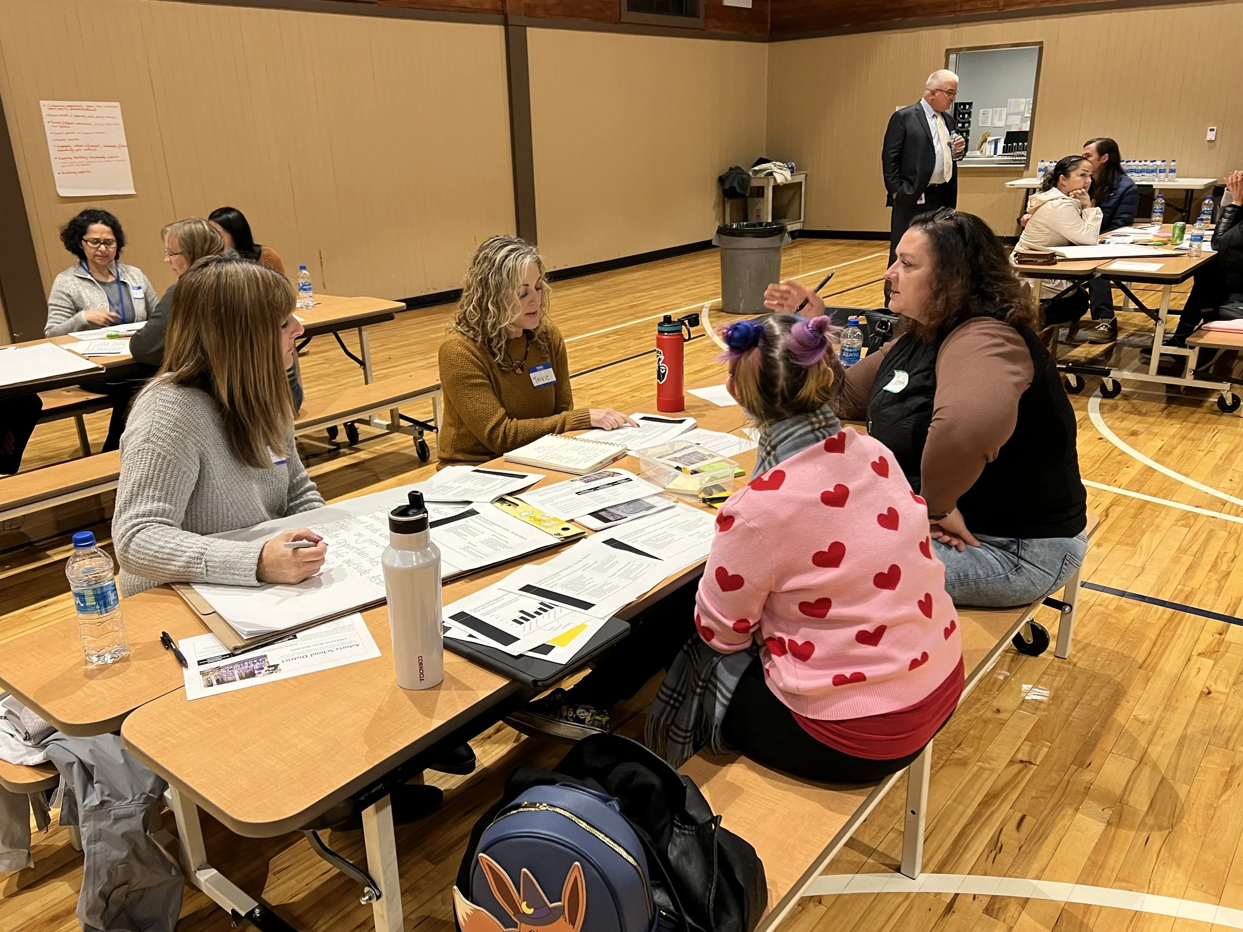 A group of people sitting around a table in a gymnasium, talking to each other, with papers on the table in front of them