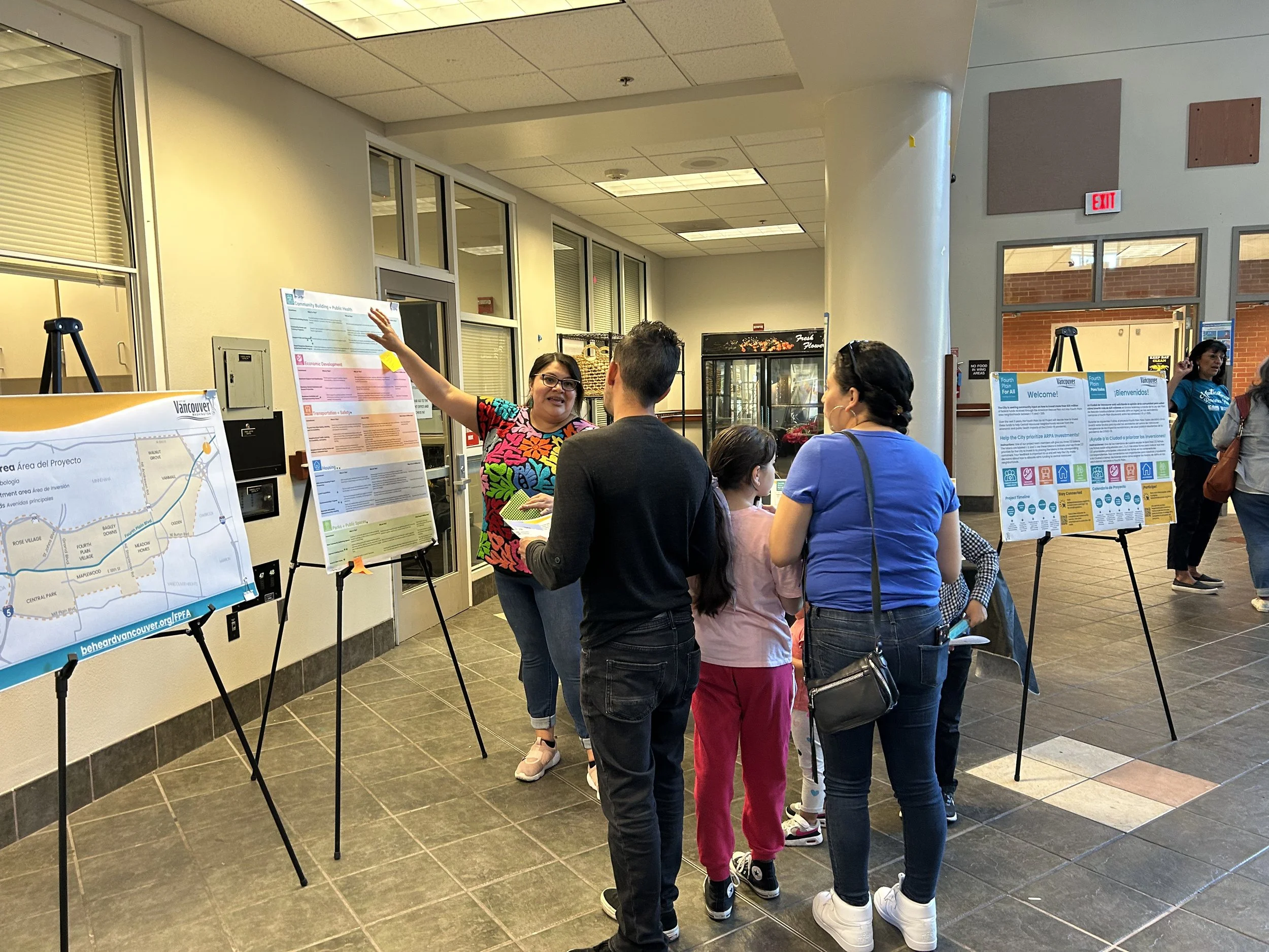 An image of a woman in a colorful shirt and glasses pointing to a display board engaging with a small group of people