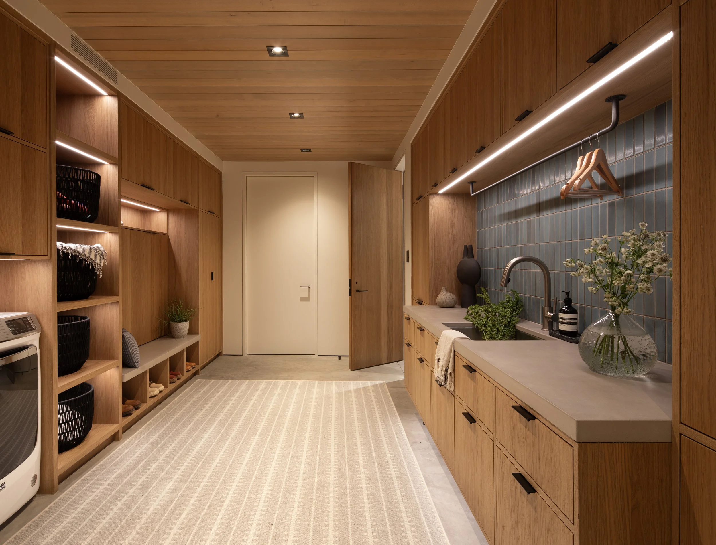 A modern laundry room with wooden cabinets, a sink, and a plant on the countertop. Shelves on the left hold baskets and shoes. Overhead are warm wood panels on the ceiling with recessed lighting.
