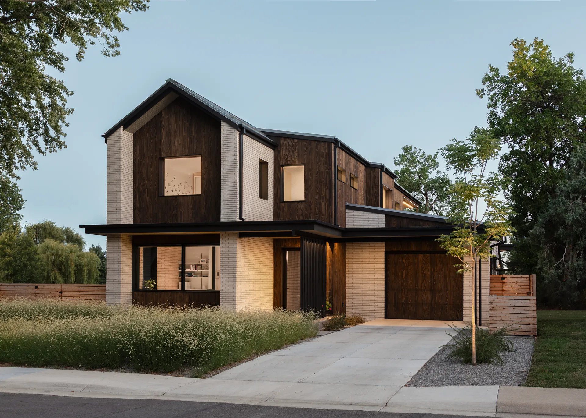 Modern two-story house with a combination of white brick and dark wood exterior, surrounded by trees and a well-maintained yard. Large windows reveal a cozy interior, and the driveway leads to a garage.