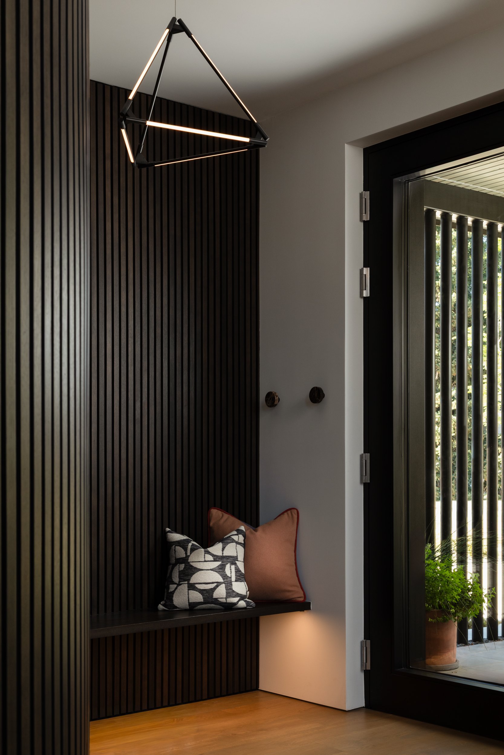 A cozy seating nook with dark wooden paneling, two pillows with black and white geometric and orange designs, located near a glass door with black frame, overlooking a garden with greenery.