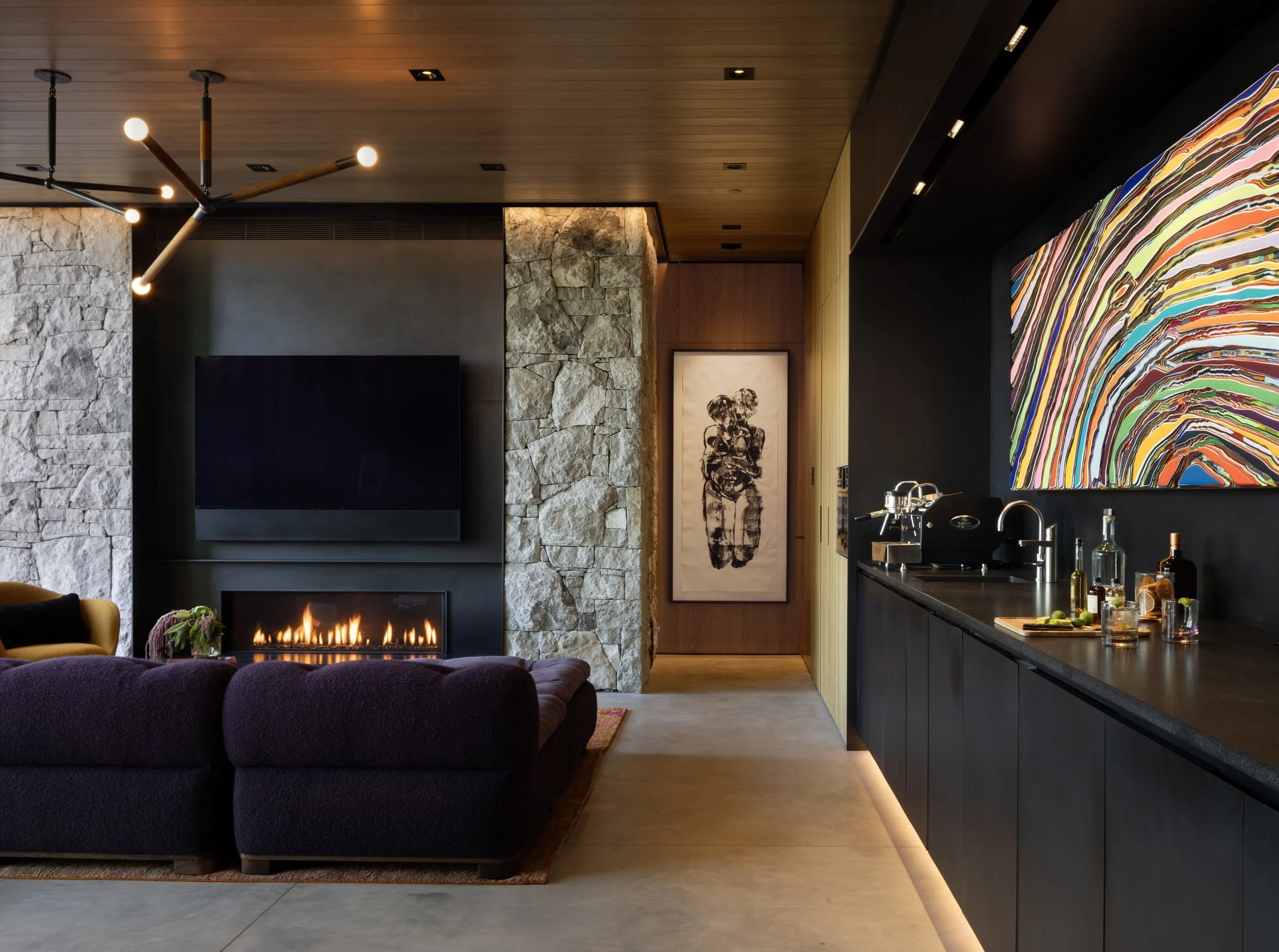 A modern living room with a stone fireplace, flat screen TV, abstract artwork, wooden ceiling, and a dark kitchen counter with a sink and bottles.