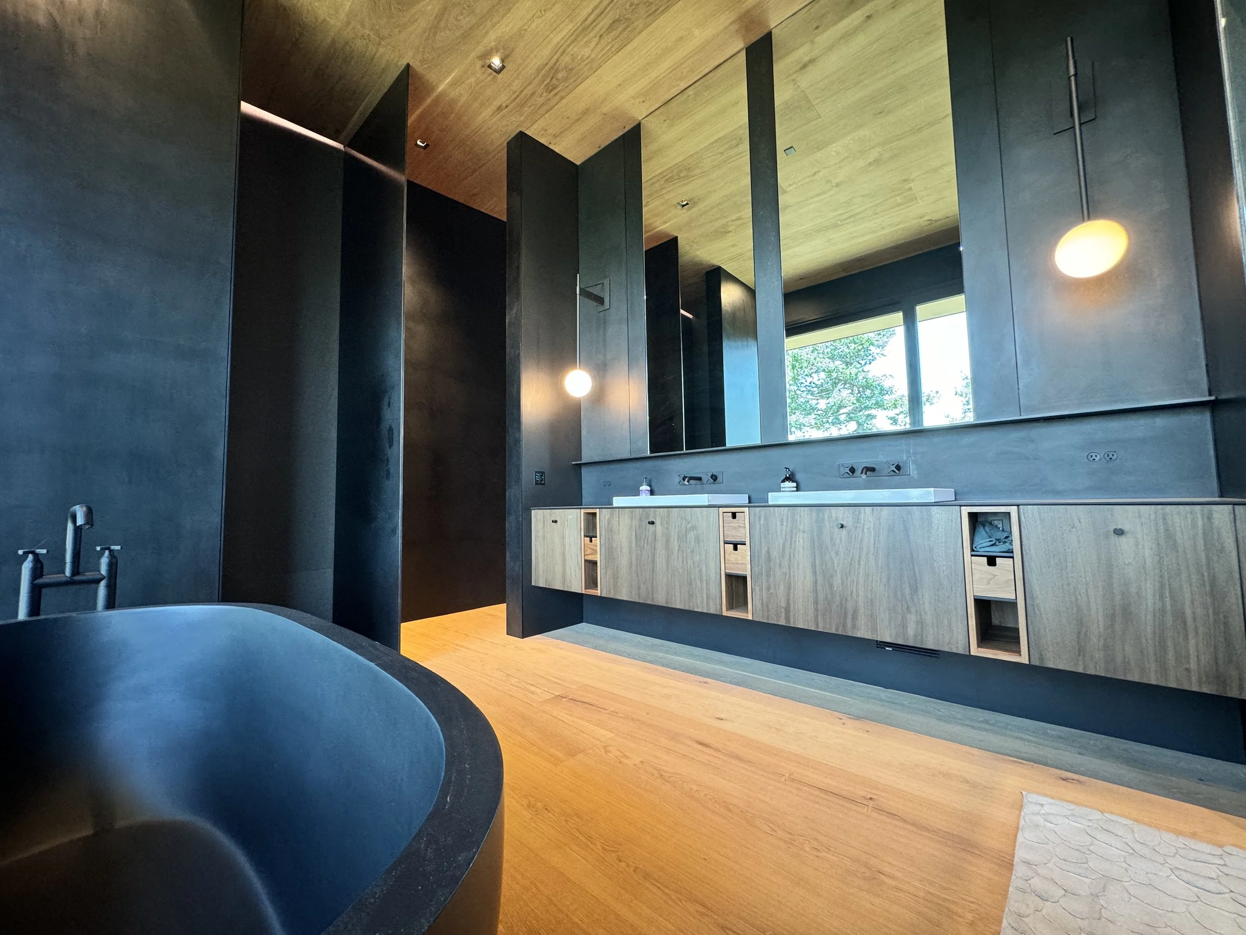 Modern bathroom with black and wood accents, large mirror, and natural light from window showing trees outside.