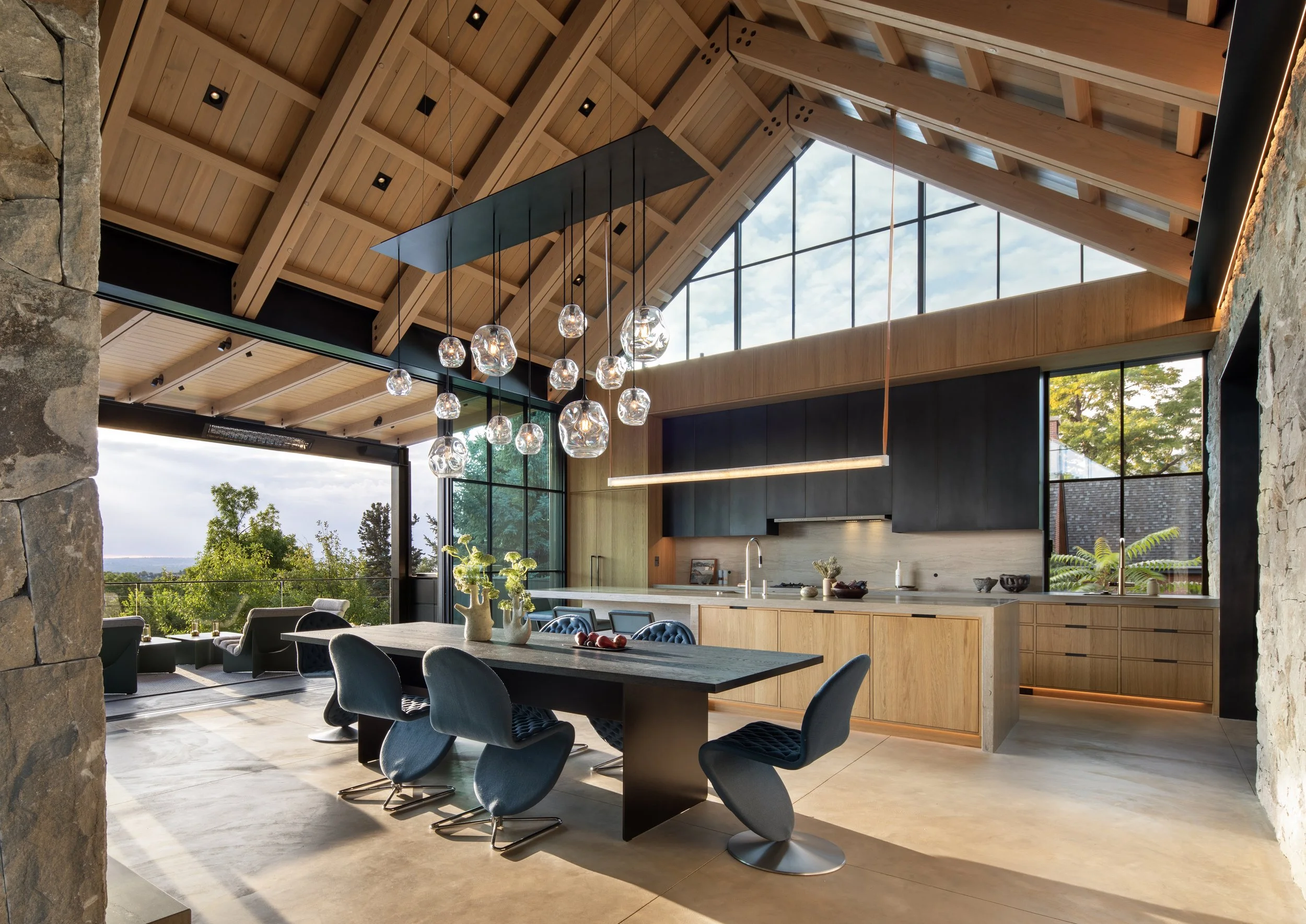 Open-concept kitchen and dining area with large windows, wooden ceiling, modern lighting fixtures, stone walls, and outdoor greenery visible through windows.