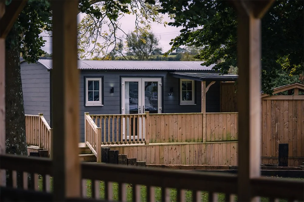 View of a small gray house with white trim through a wooden structure, surrounded by trees and a wooden fence.