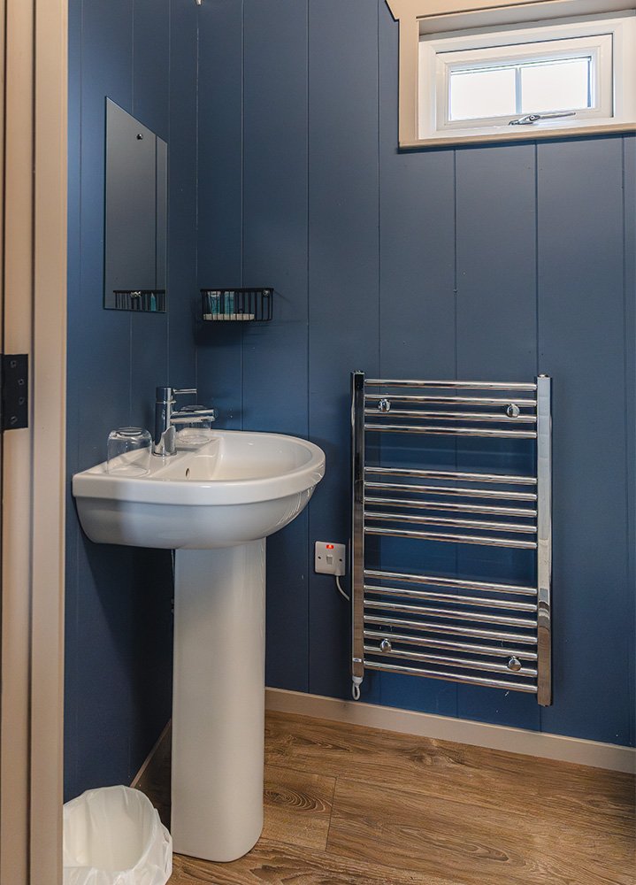 Small bathroom with blue panel walls, a white pedestal sink with glasses on it, a mirror, a small wire shelf, a window, a chrome towel radiator, a power socket, a wooden floor, and a small trash bin.