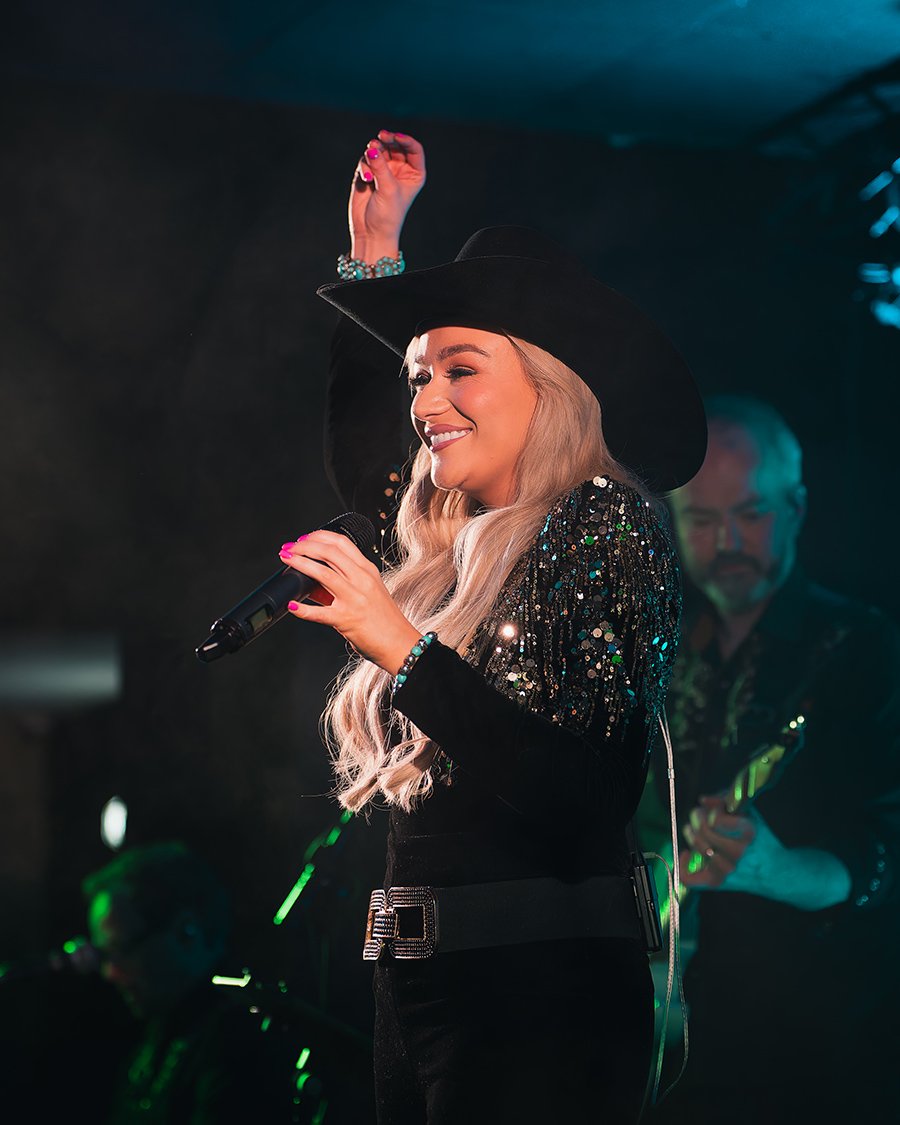 Claudia Buckley wearing a black hat, sequined jacket, and holding a microphone, smiles on stage with band members in background.