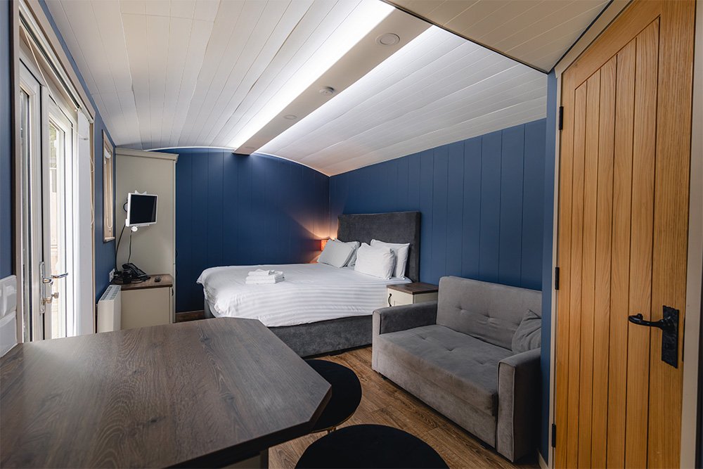 Glamping pod room with a bed, a small gray sofa, a wooden door, a wall-mounted TV, and a window, in a cosy space with a sloped ceiling and wooden flooring.