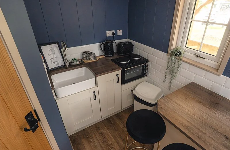 Small kitchen with blue wall panels, white cabinets, a wooden countertop, a sink, a microwave oven, a black toaster, a kettle, a window with a hanging plant, and a stool.