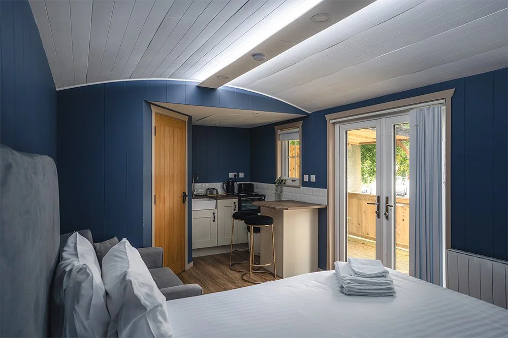 Glamping Pod with a bed, sitting area, small kitchenette, and a glass door leading to an outdoor patio, in a blue and white color scheme.