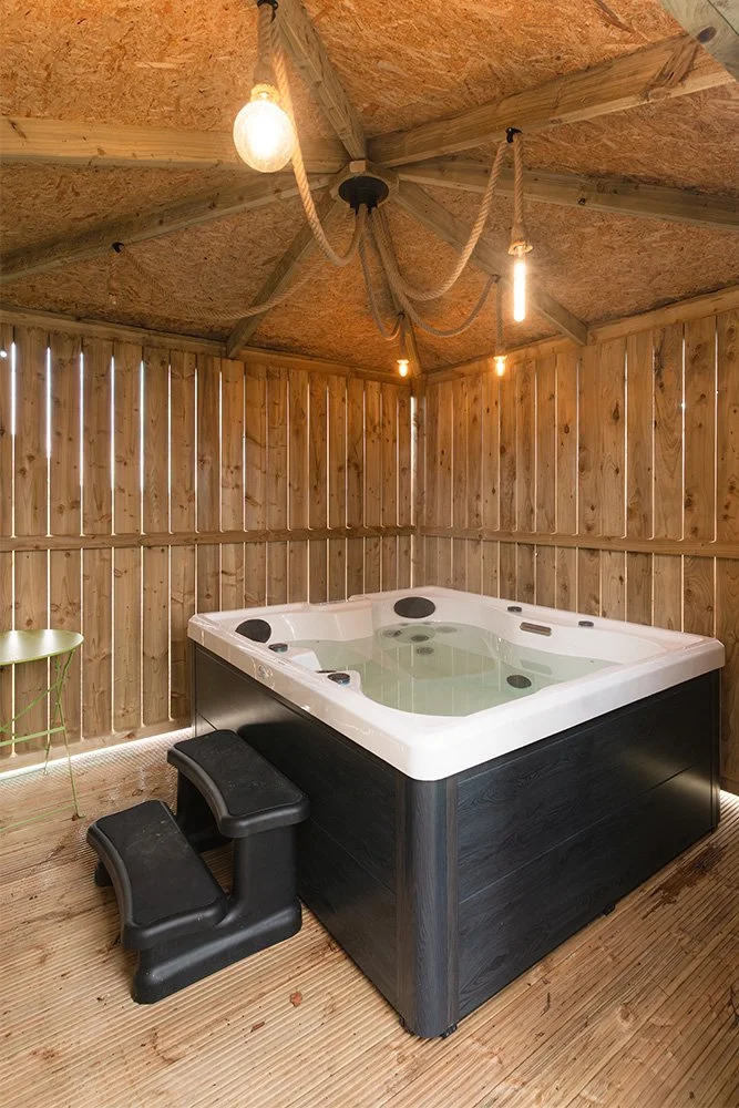 Wooden fenced outdoor hot tub with black exterior, black steps, and a wooden floor inside a rustic wooden shed or gazebo.