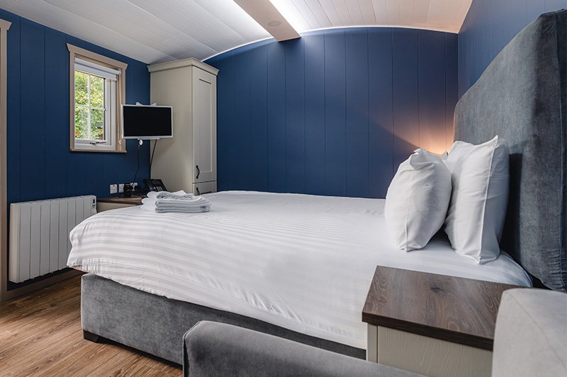 A glamping pod room with a bed, two pillows, a folded towel, a small window, a wall-mounted TV, a white wardrobe, and a blue accent wall.