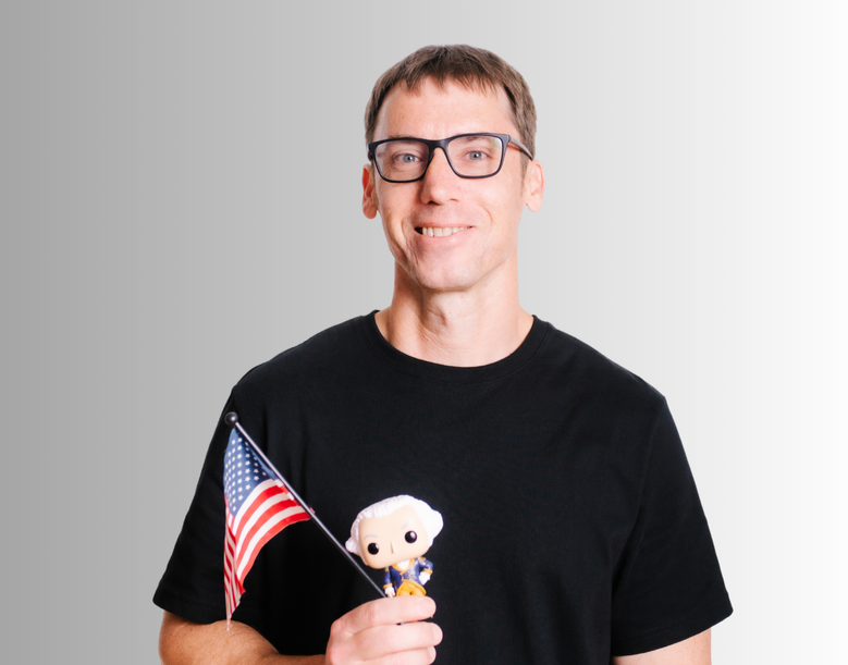 Man with glasses holding an American flag and a small plush toy of a historical figure, smiling against a neutral gray background.