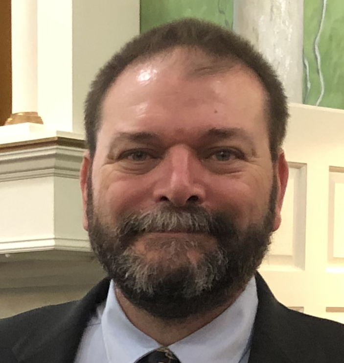 Close-up of a smiling man with a beard, wearing a suit and tie, indoors with a decorative background.