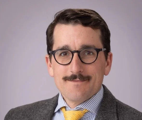 A man with dark hair, glasses, a mustache, wearing a gray suit, a striped shirt, and a yellow tie, posing against a plain light background.
