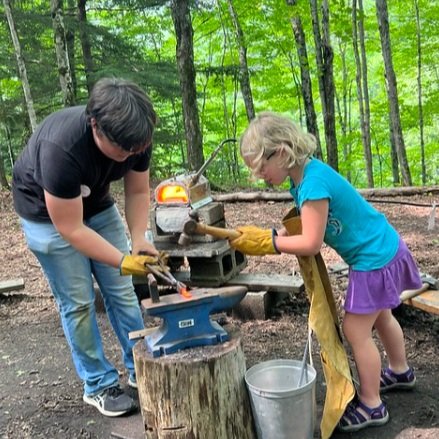 An adult and child working with hot metal on an anvil in the outdoors