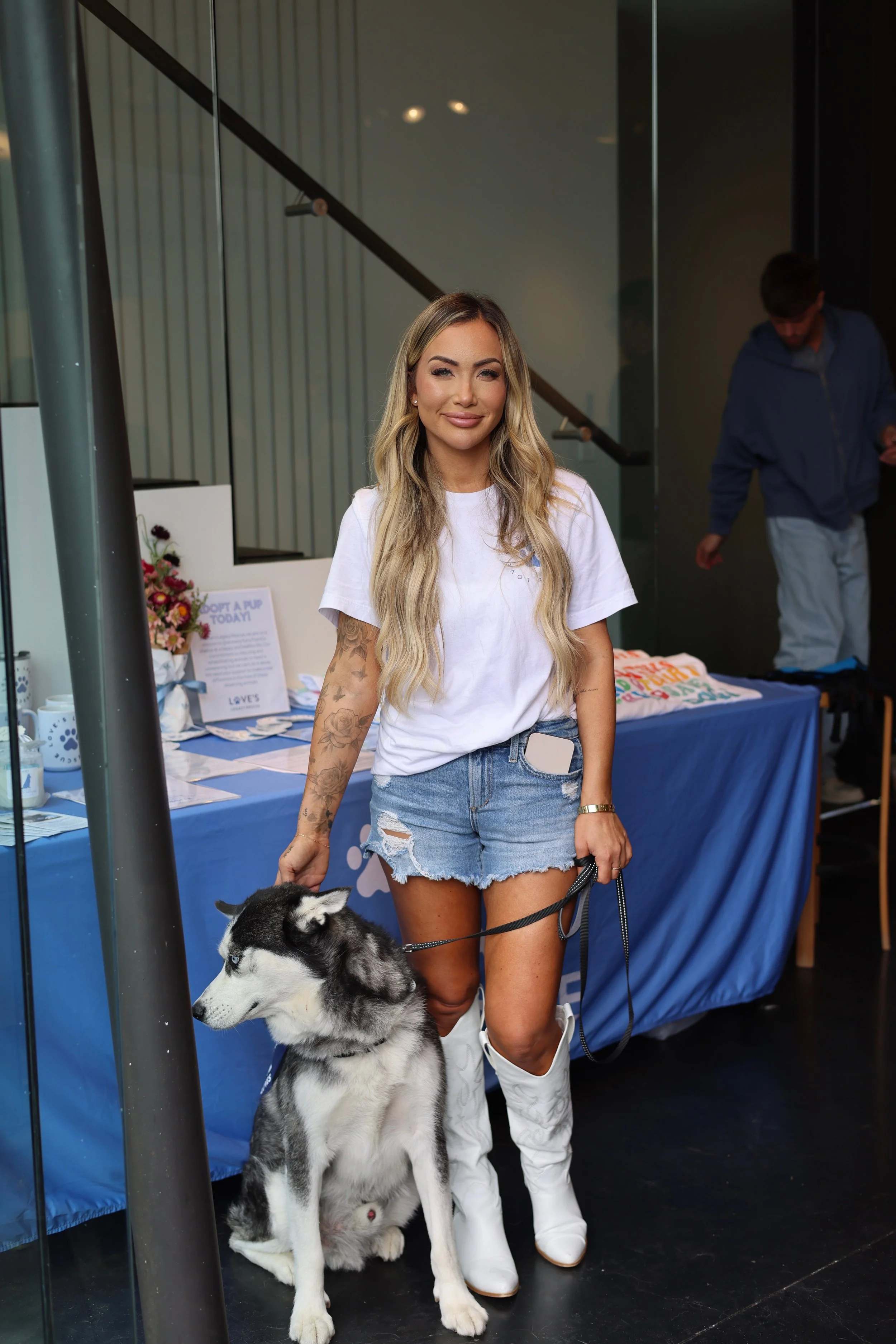 A woman with long blonde hair, wearing a white t-shirt, denim shorts, and white boots, standing next to a husky dog at an indoor event or booth, with a person in the background and a table with informational items and flowers.