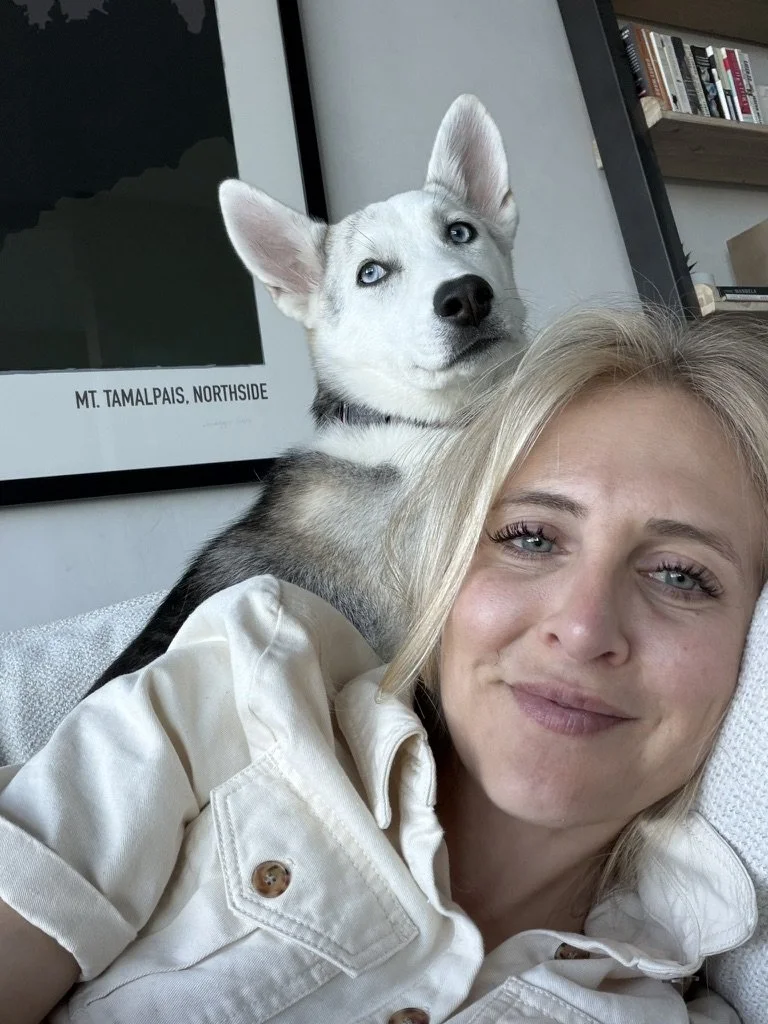 A woman lying on a couch with a Siberian Husky puppy resting on her shoulder. The woman is smiling softly, and the puppy has striking blue eyes with an alert expression. In the background, there is a framed poster of Mount Tamalpais, Northside, and books on a shelf.