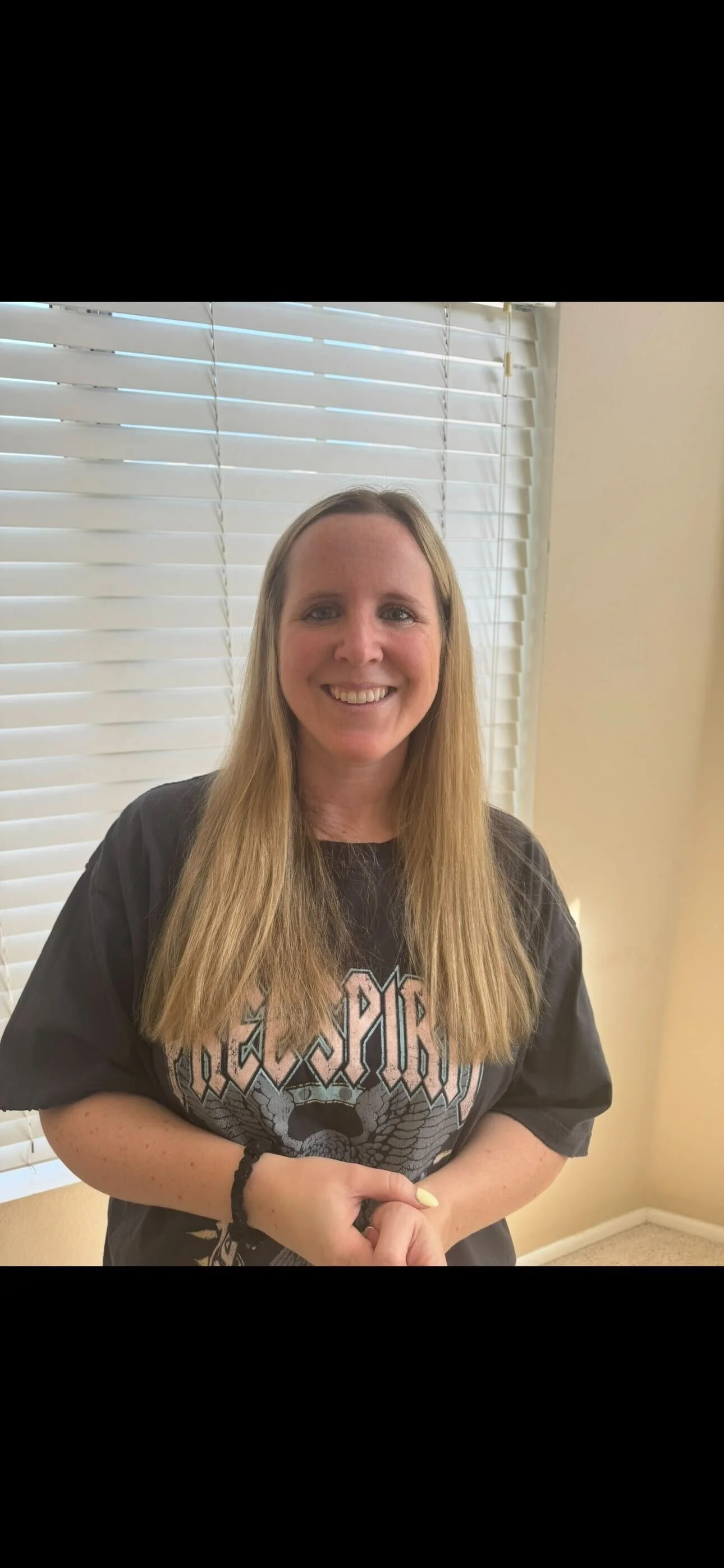 A smiling woman with long blonde hair standing in front of white window blinds, wearing a black shirt with an 'AC/DC' logo.